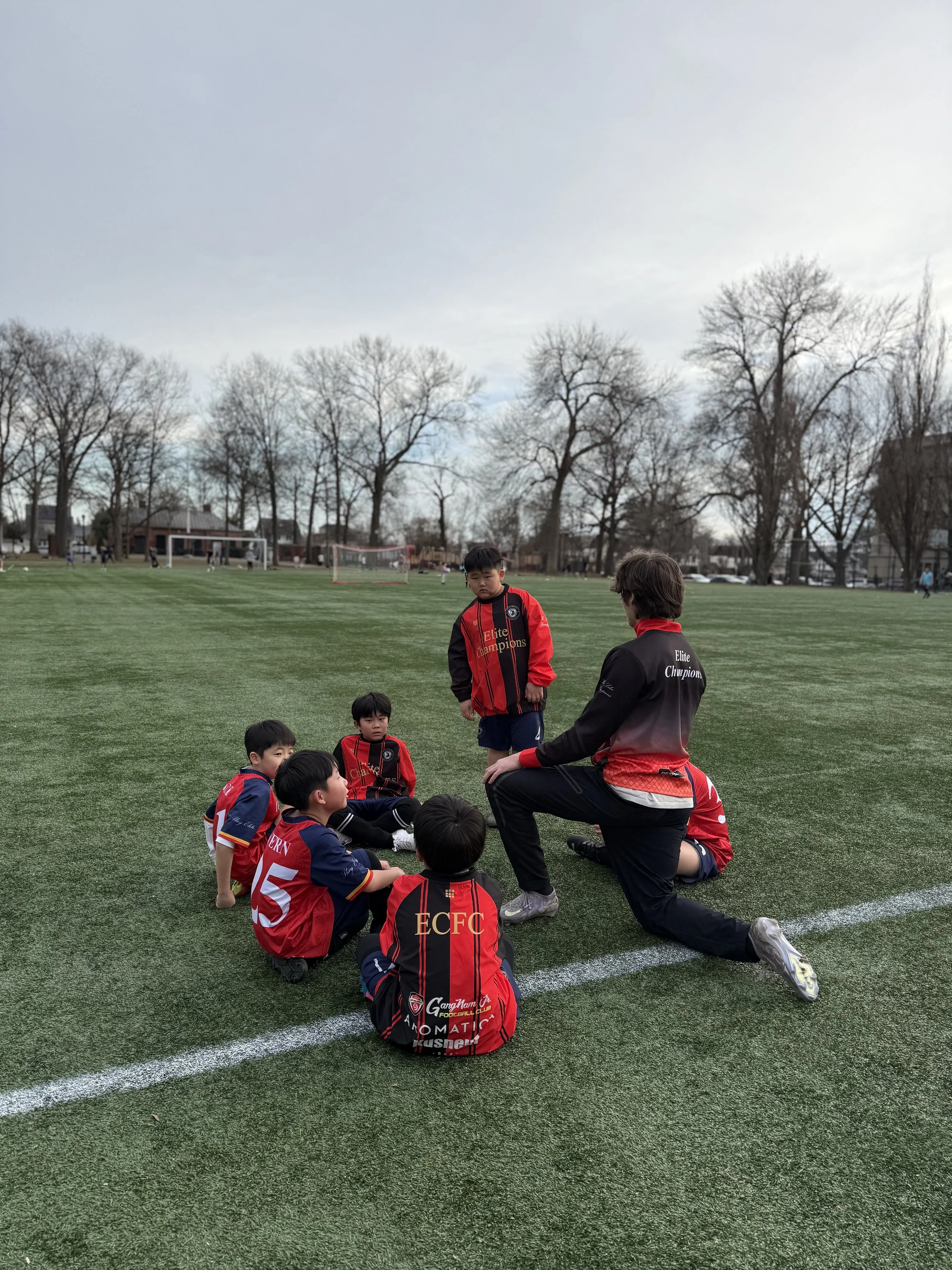 A group of young boys and a coach on a football field, some boys are sitting while the coach is kneeling and speaking to them, all wearing red and black football uniforms. Trees and a goalpost are visible in the background.