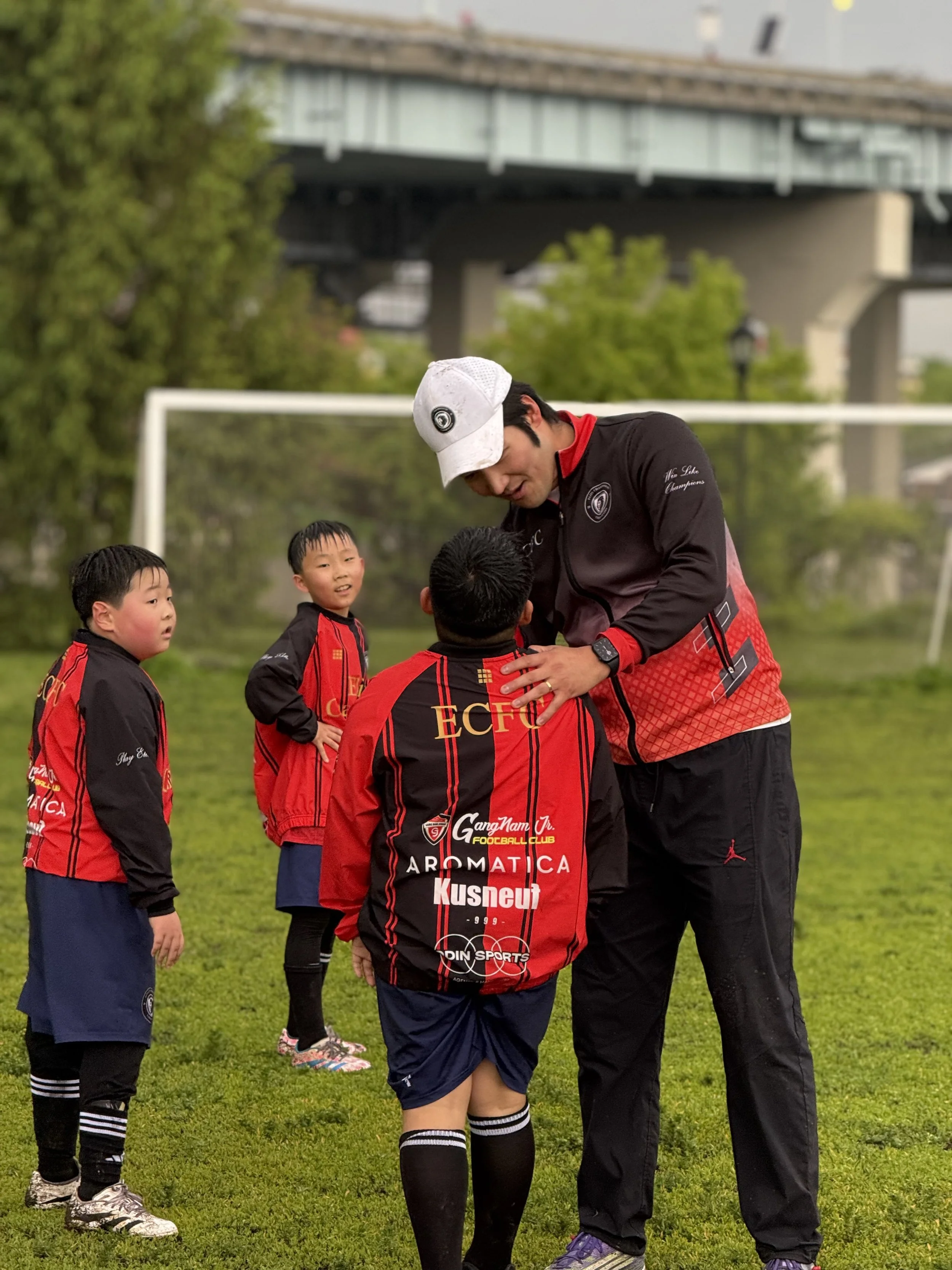 A group of young boys in soccer uniforms standing on a grassy field, listening to an adult coach who is giving instructions. The boys are wearing black and red jerseys, and the coach is wearing a black jacket with a white cap. There is a soccer goal and trees in the background.