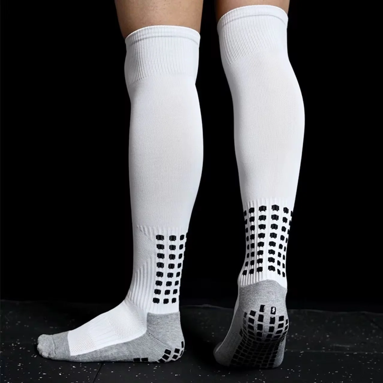 White athletic compression socks with black dotted and grid patterns, gray toe and heel sections, worn by a person standing against a black background.