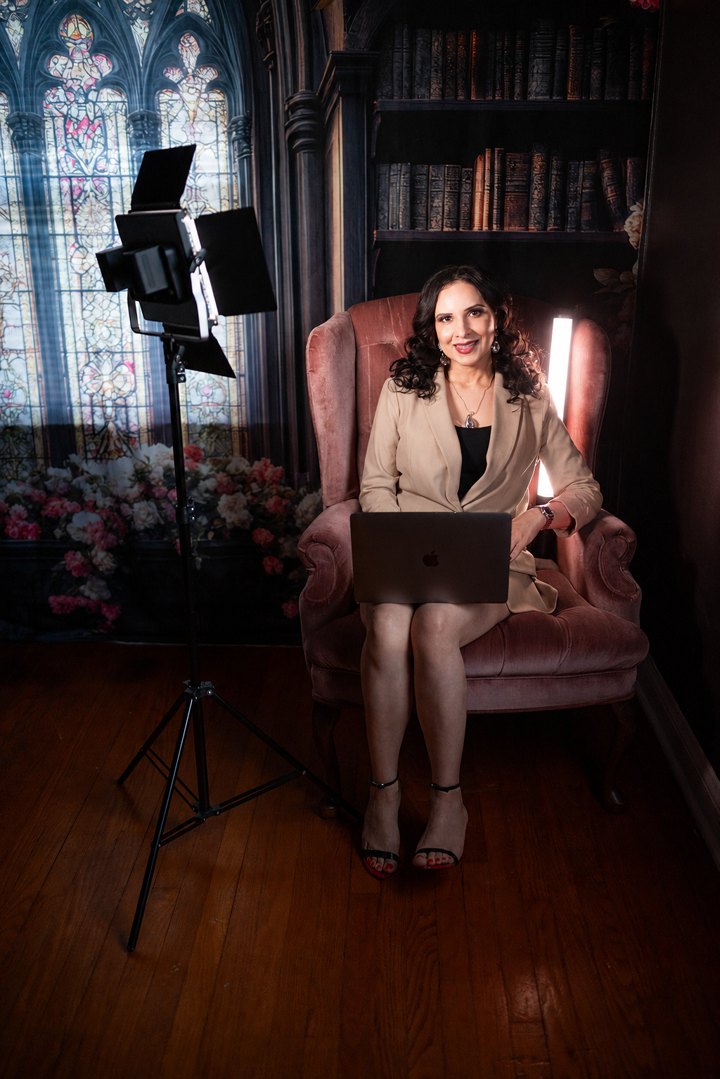 A woman sitting on a vintage armchair in a dimly lit room, with a laptop on her lap, a camera and light stand nearby, a dark wooden bookshelf behind her, and a stained glass window in the background.