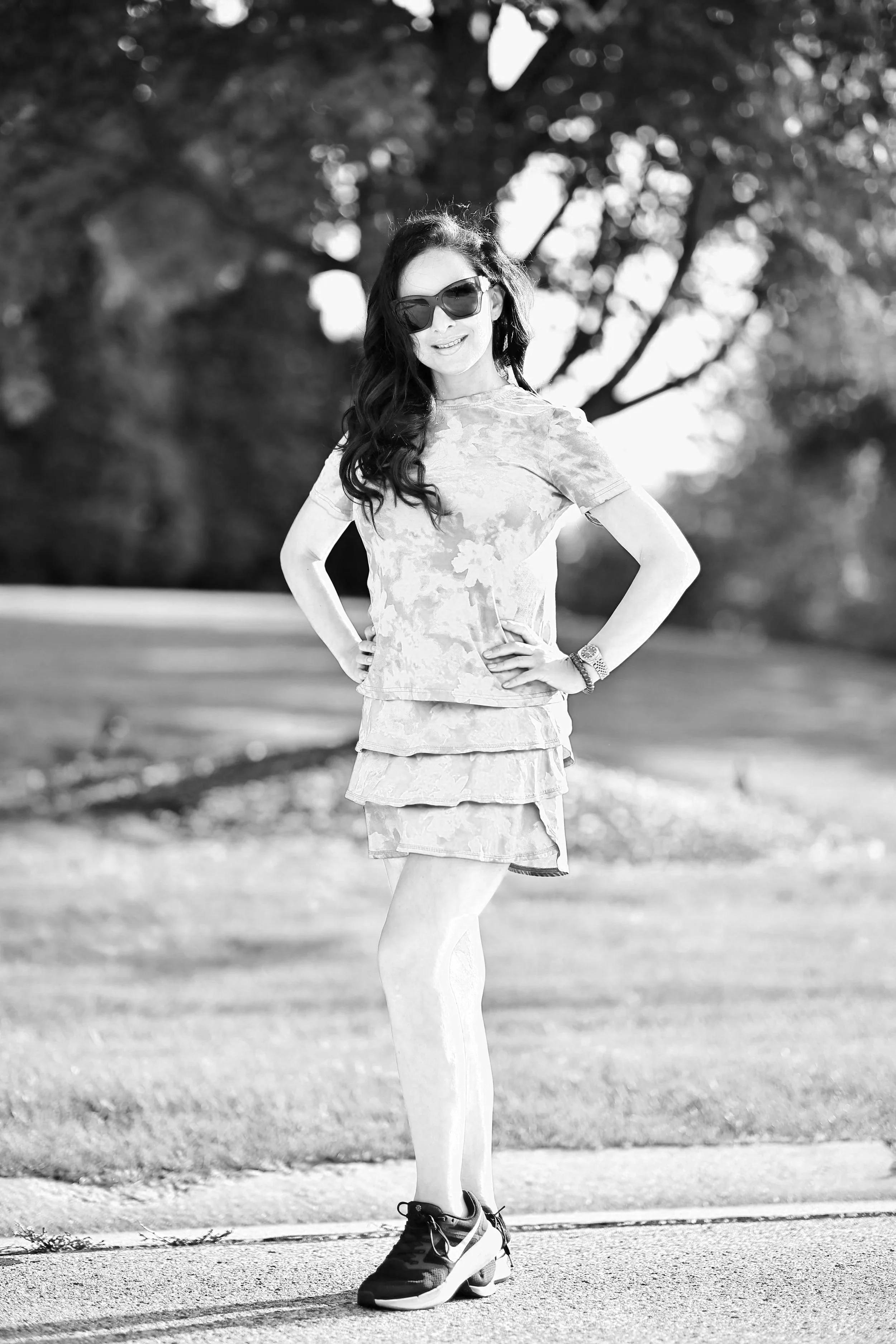 A smiling woman with long dark hair wearing sunglasses, a patterned top, a skirt, patterned tights, and sneakers, standing outdoors with her hands on her hips against a background of trees and grass.