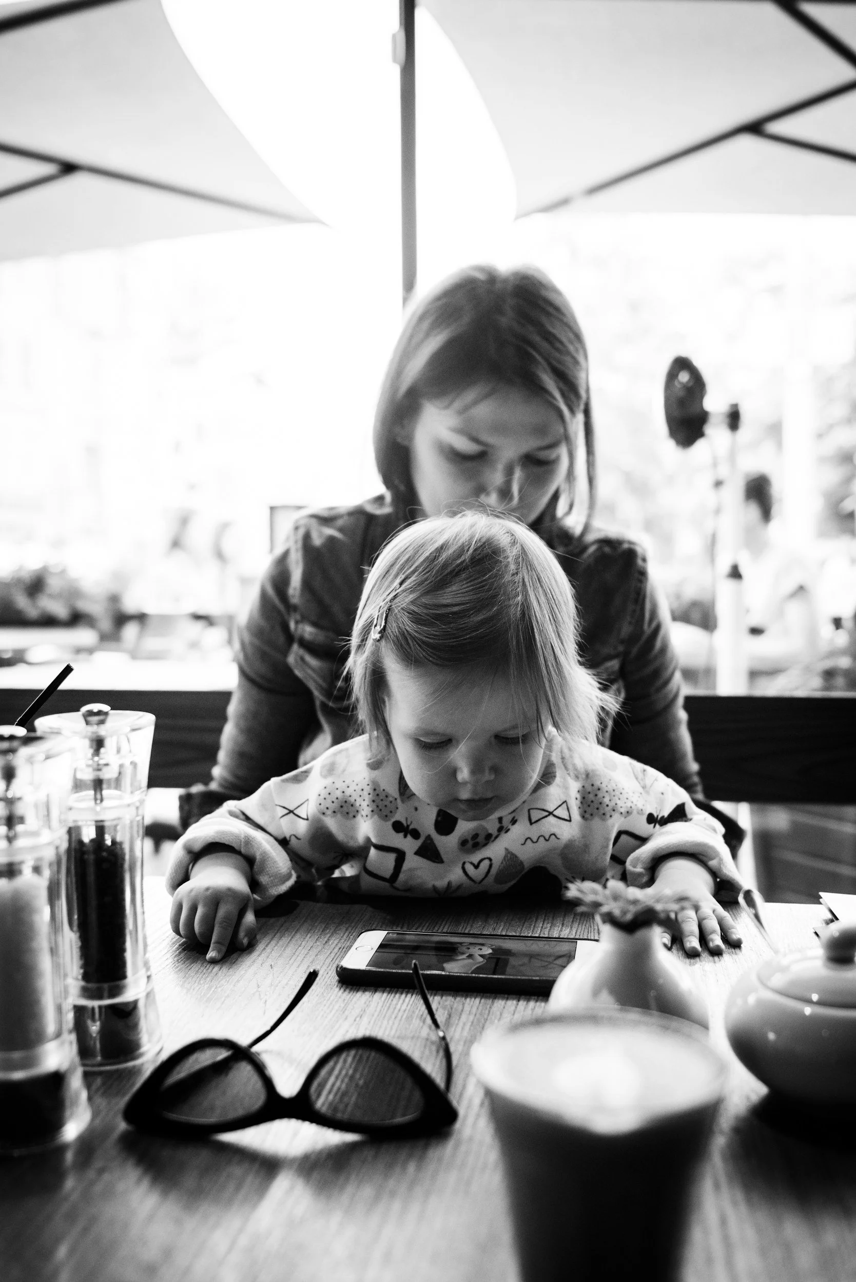 A woman and a young girl are sitting at a table in a restaurant or cafe, looking at a smartphone. The woman is behind the girl, leaning forward and looking down. There are umbrellas outside the window, and various objects on the table, including glasses, a bowl, and what appears to be a tablet or another phone.