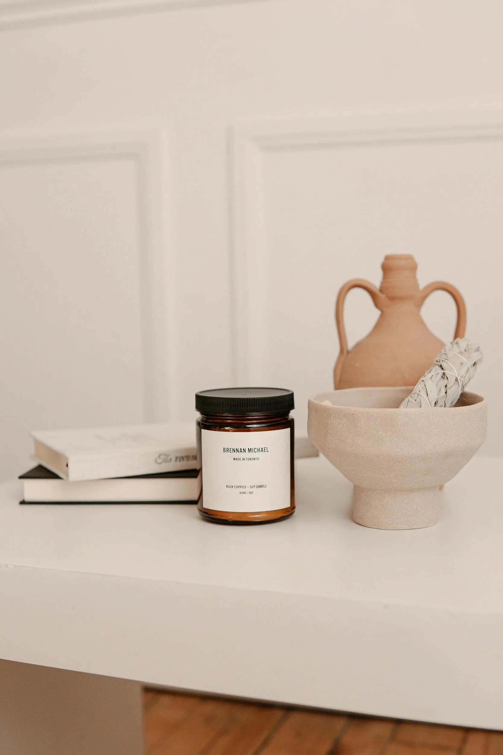 A white surface with a small collection of decorative objects including a ceramic vase, a scented candle, a small bowl, and a stack of books.