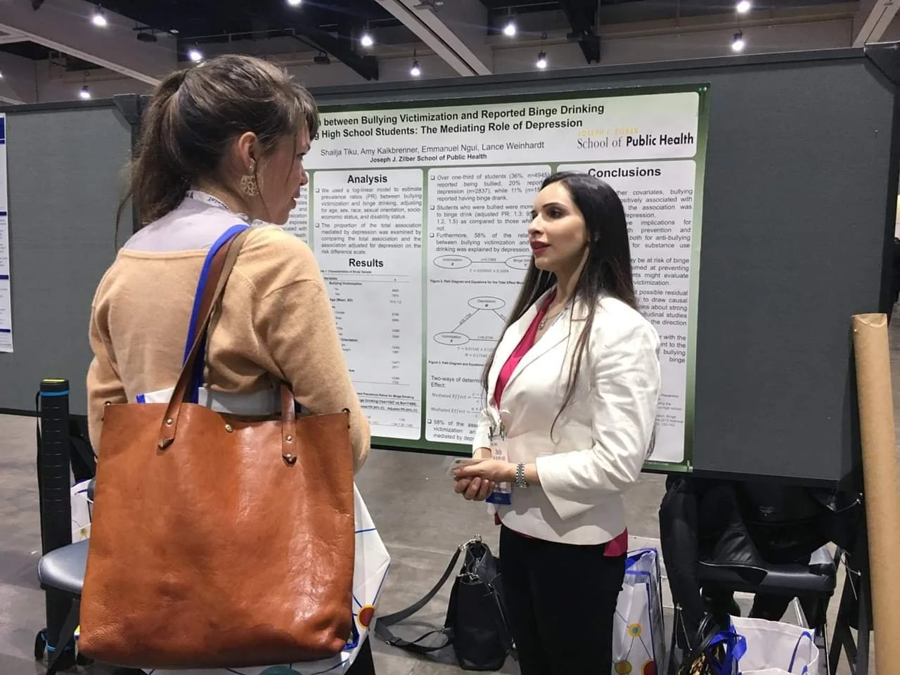 Two women talking in front of a scientific poster at a conference or academic event. One woman has long dark hair, wearing a white blazer and pink top. The other has brown hair tied back, wearing a beige sweater with a large brown leather tote bag.