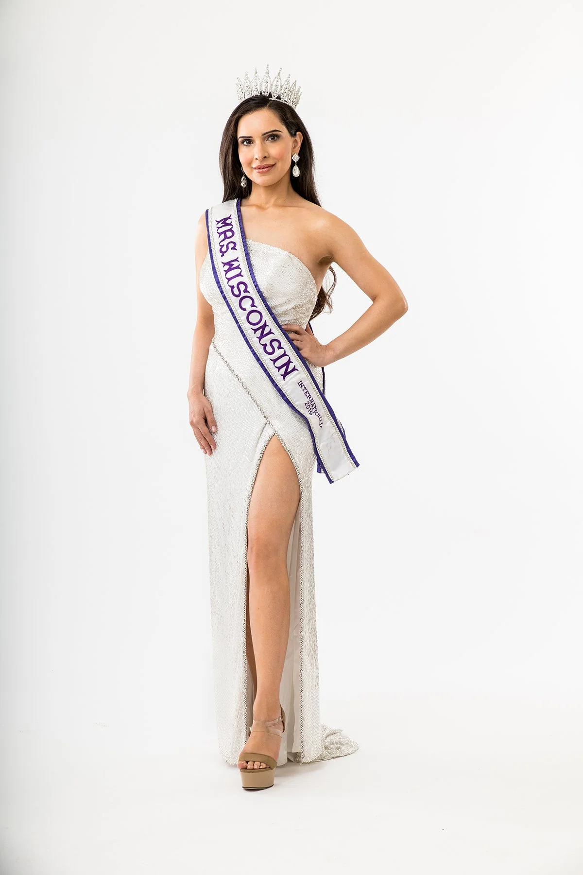 Beauty queen wearing a tiara and sash reading 'MISS NY USA 2023' in a long, white, high-slit gown with heels, posing against a white background.