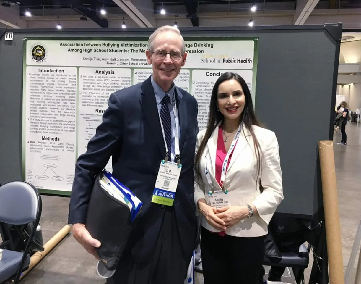 Two professionally dressed individuals, a man and a woman, standing side by side at a conference, smiling, with a research poster behind them. The man is holding a folder and has a conference badge, and the woman's badge indicates she is a speaker.