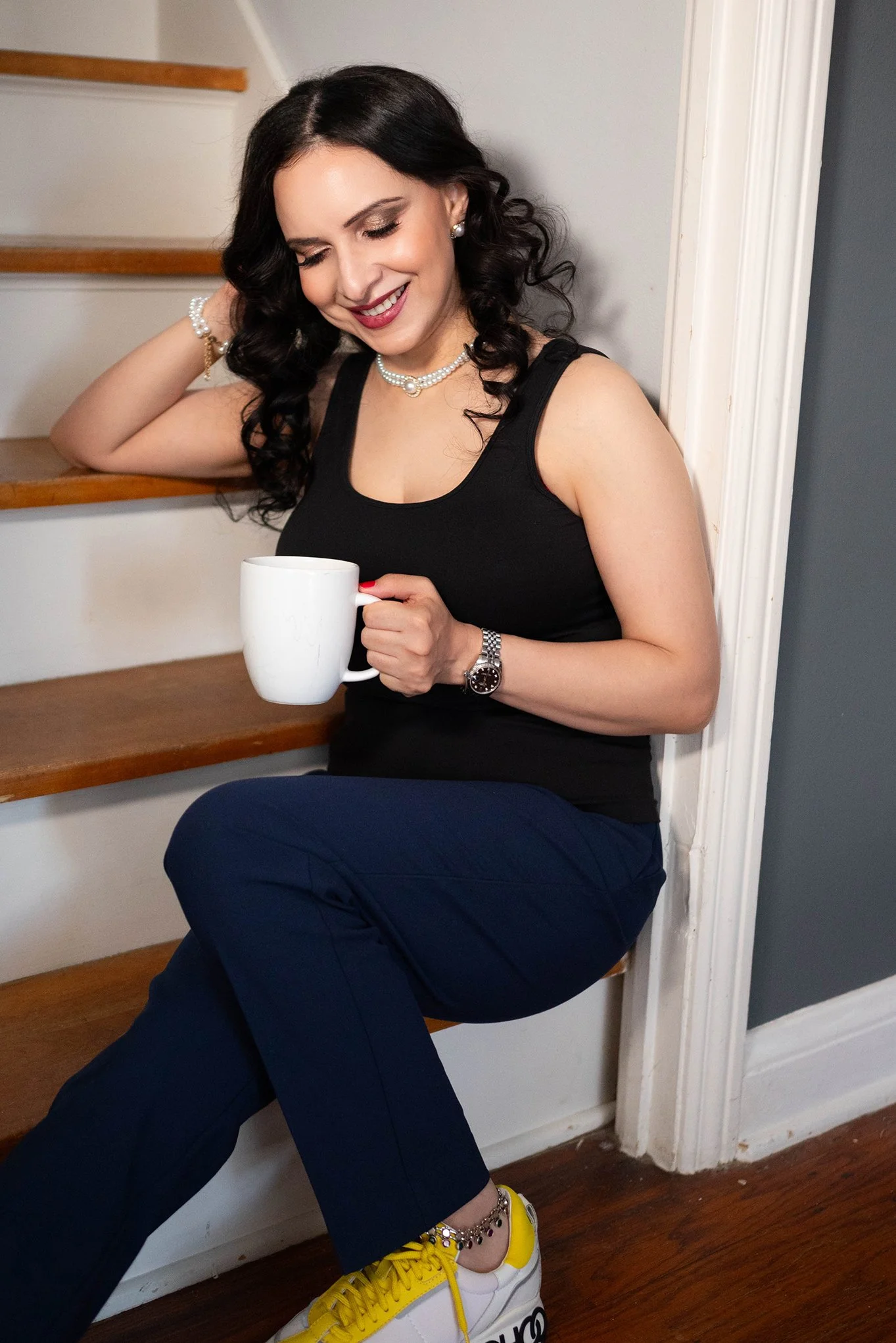 A woman with long black curly hair wearing a black tank top, pearl jewelry, and a watch, smiling and holding a white mug, sitting on a staircase with wooden steps and white walls.