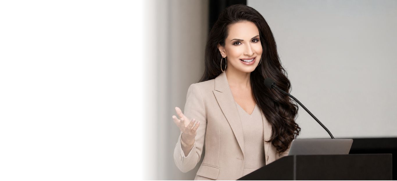 A woman with long dark hair, wearing a beige blazer, speaking at a podium with a microphone, smiling and gesturing with her right hand.