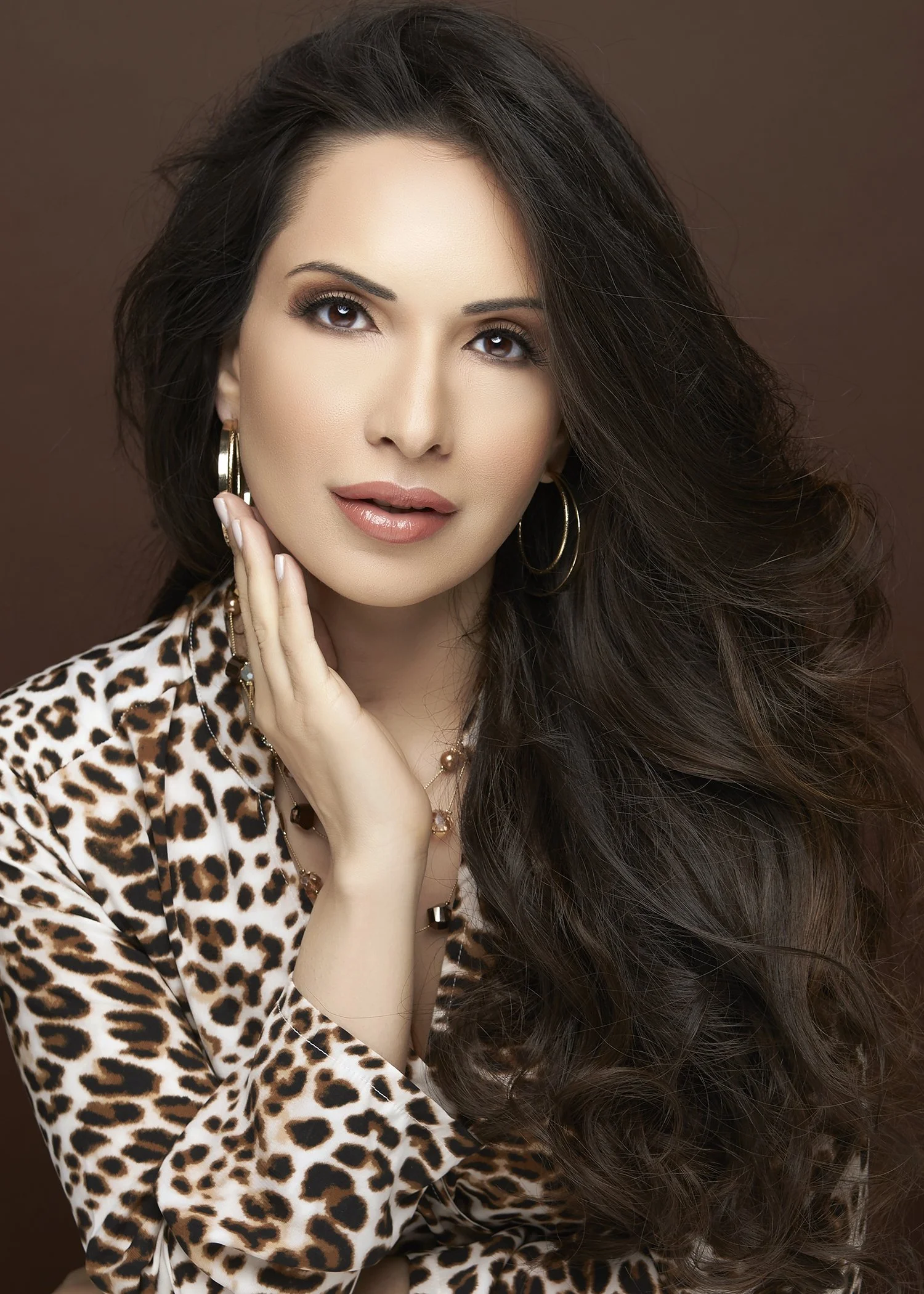 A woman with long, wavy dark brown hair, wearing gold hoop earrings, a leopard print top, and jewelry, against a plain brown background.