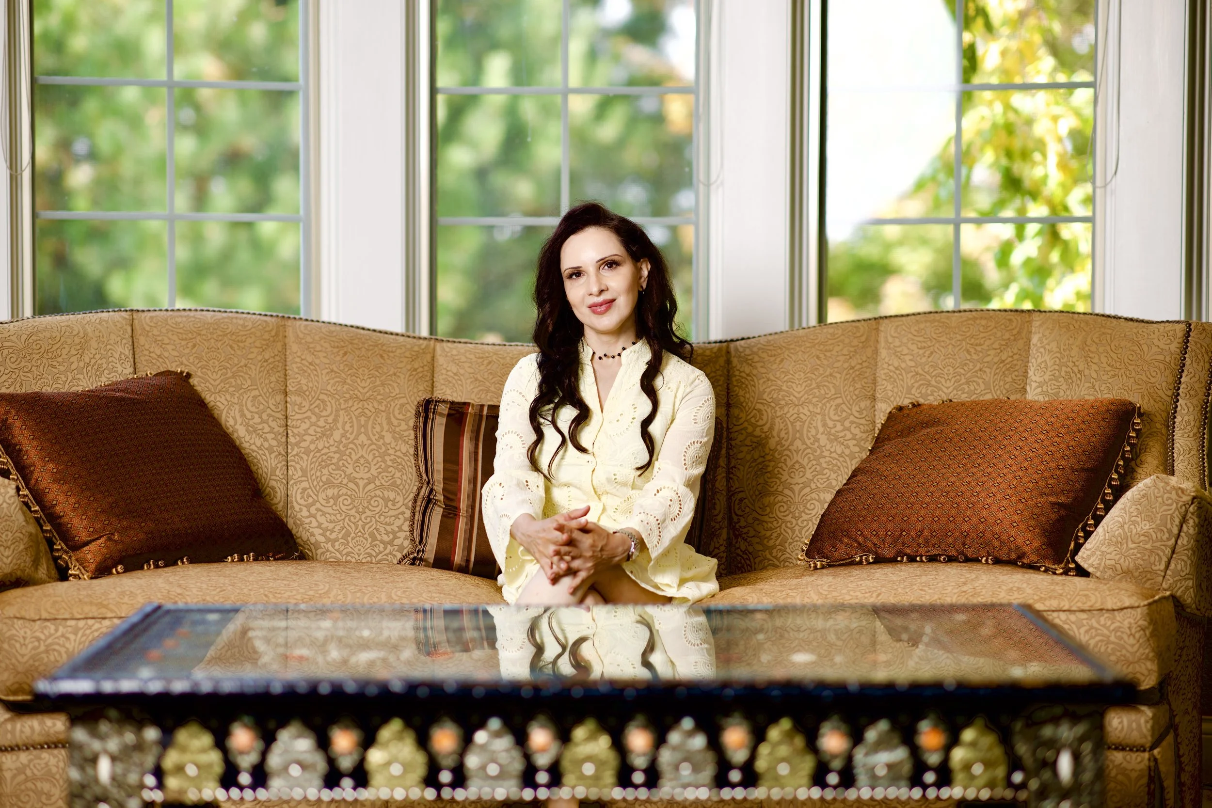 A woman with long dark hair sitting on a beige patterned couch in front of large windows with green trees outside, wearing a light yellow dress with lace details and a beaded necklace.
