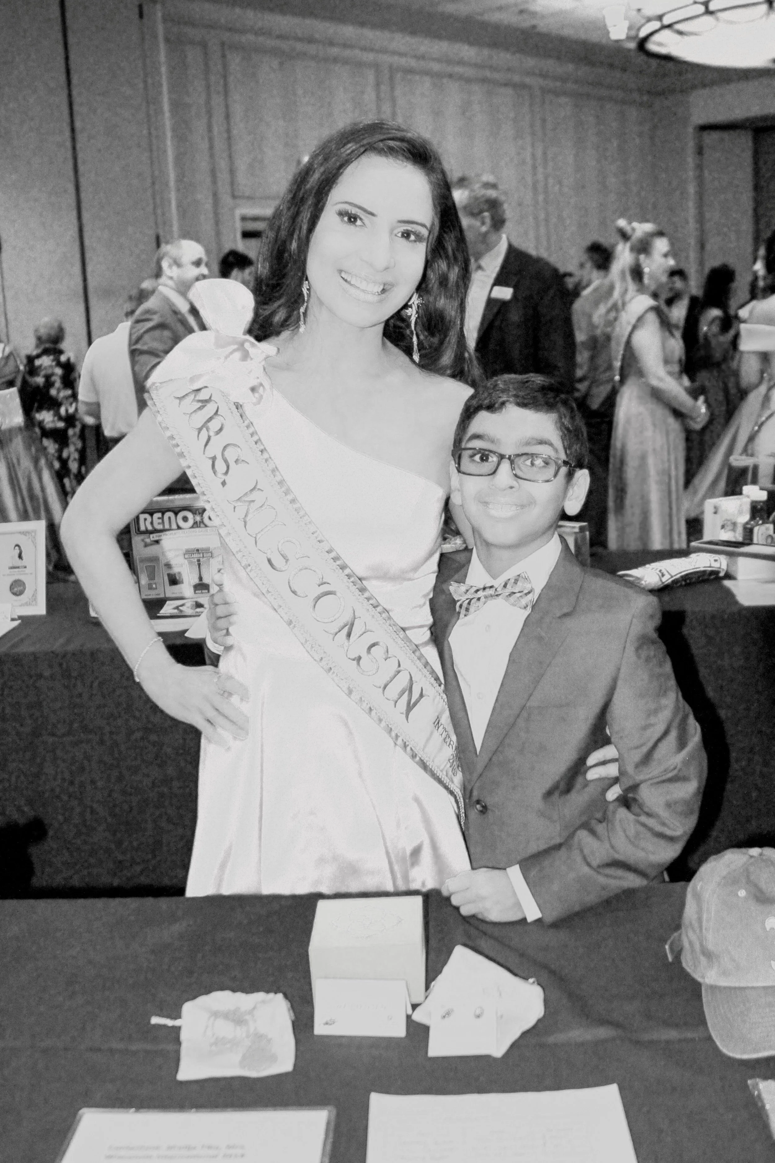A woman wearing a sash that reads 'Miss Sokal' and a formal gown, standing next to a young man in a suit with glasses, at a formal event or pageant. They are in a room with other people in the background.