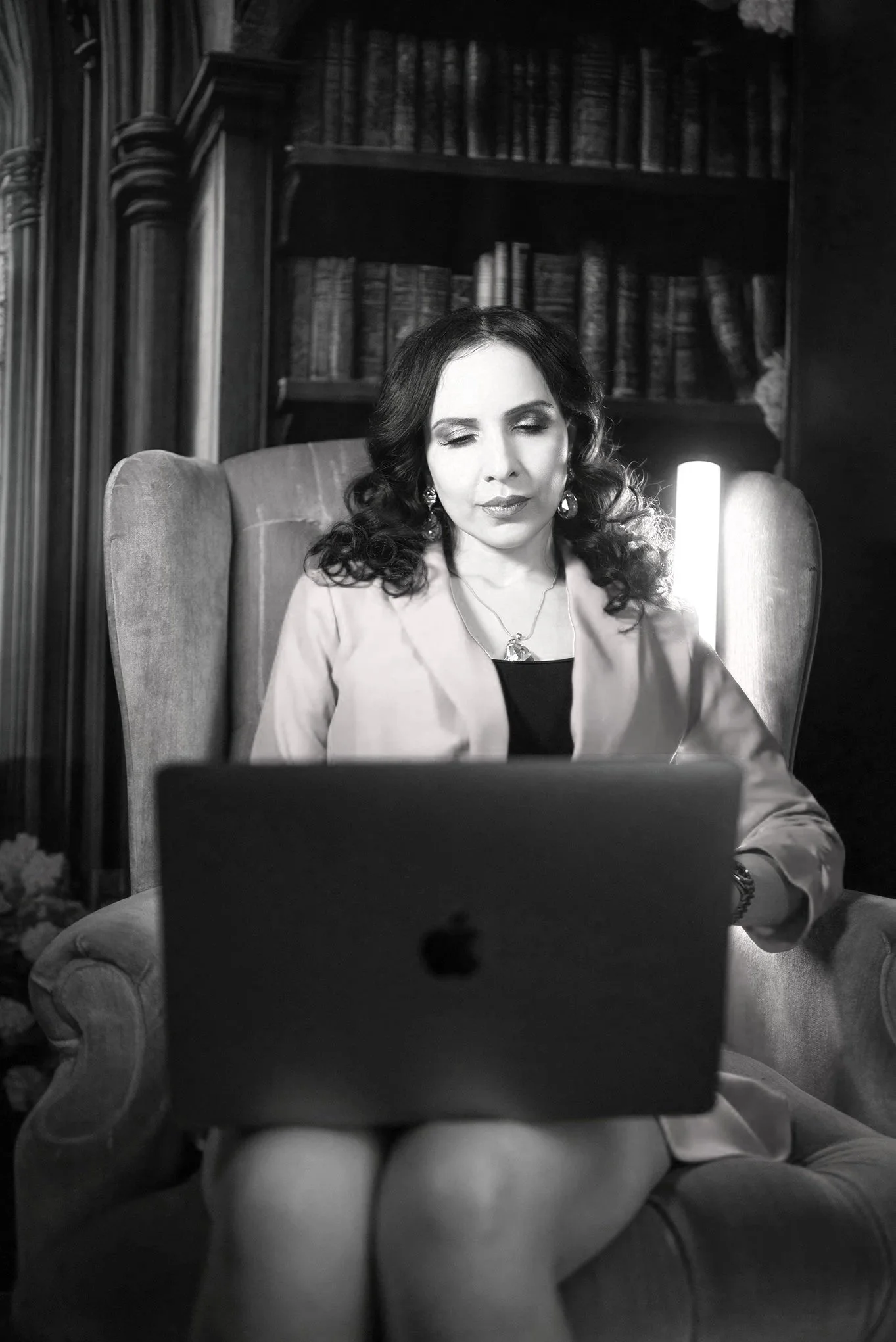 A woman with dark, wavy hair and earrings, sitting on a vintage armchair in front of a bookshelf. She appears to be reading or looking at a screen in front of her, with her eyes partially closed. The setting is a room with wooden furniture and a lamp emitting light in the background.