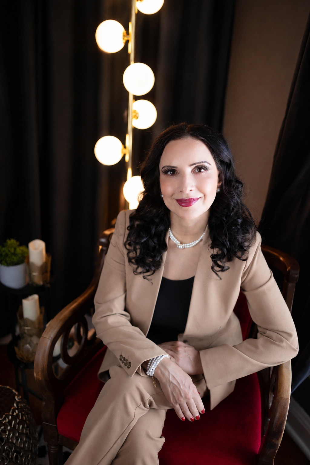 A woman with dark curly hair wearing a beige suit, black top, pearl necklace, and bracelet, sitting on a red velvet chair in a room with black curtains and warm globe lighting.