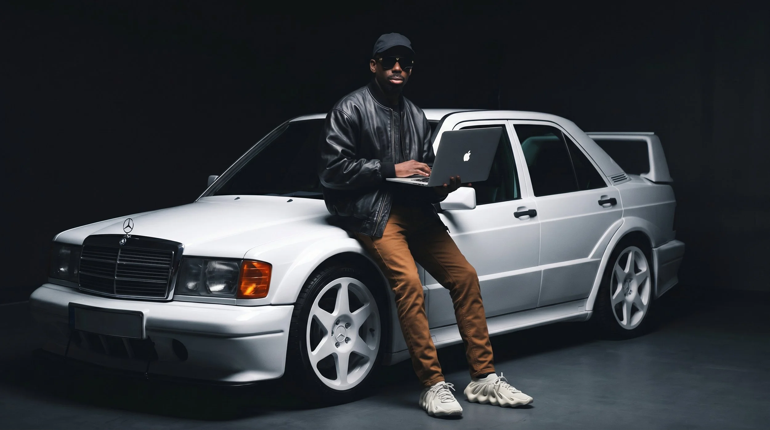 A man wearing a black leather jacket, tan pants, and white sneakers is sitting on the hood of a white vintage Mercedes-Benz car. He is holding a silver laptop and is wearing sunglasses and a black cap. The background is dark.