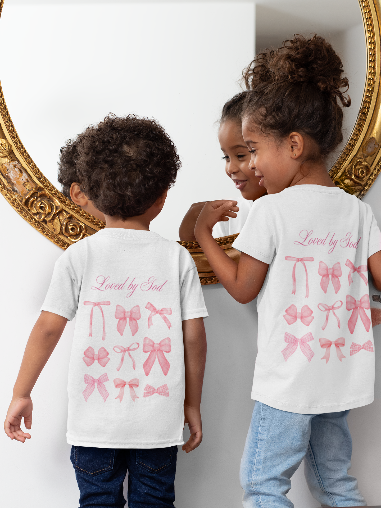 Three children standing in front of a mirror, wearing matching white T-shirts with pink bows and the phrase 'Loved by God.' The children are looking at their reflections with smiles and engaging with each other.
