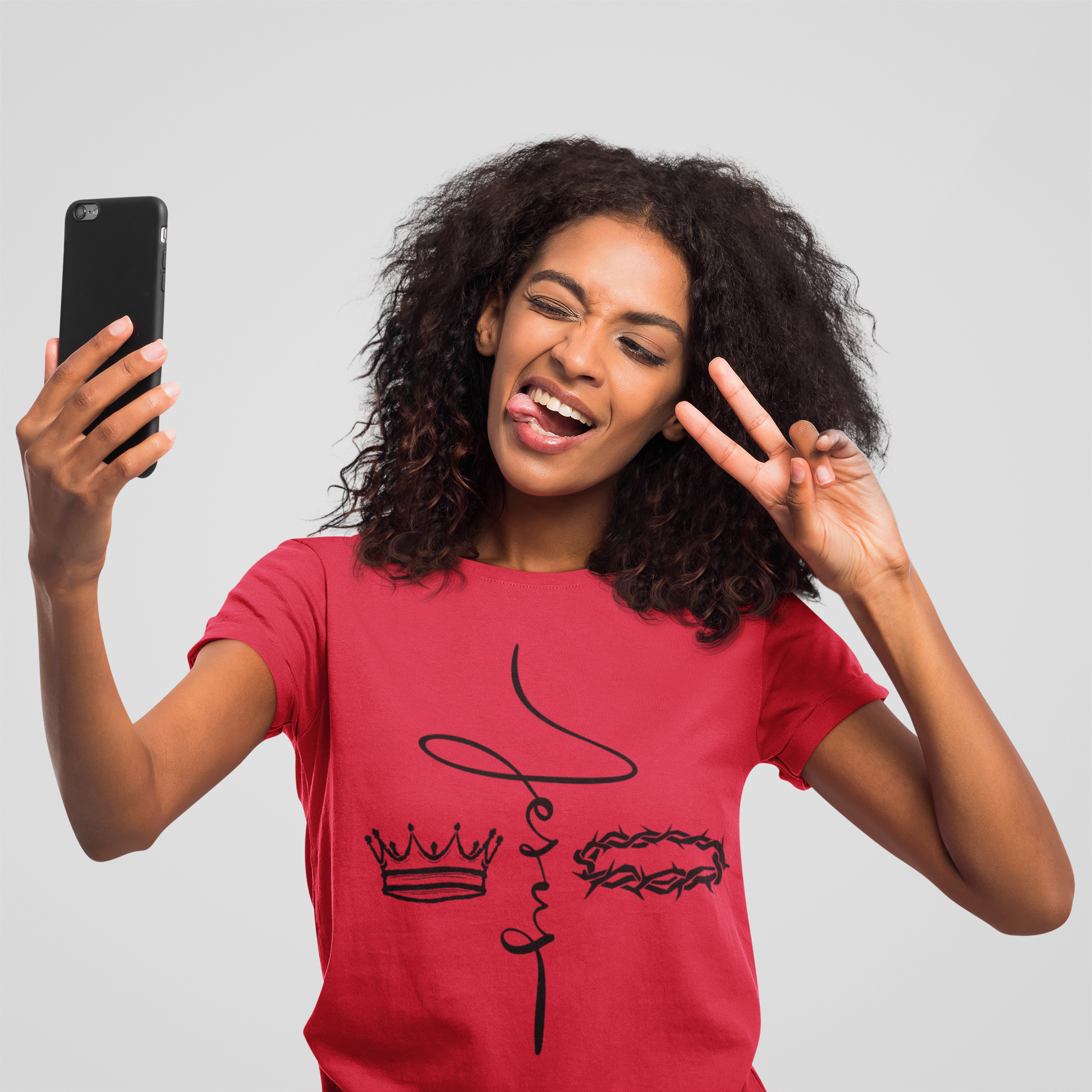 Young woman with curly hair taking a selfie, making a peace sign with her right hand, sticking out her tongue, and smiling with her eyes closed, wearing a red T-shirt with black line art graphics.