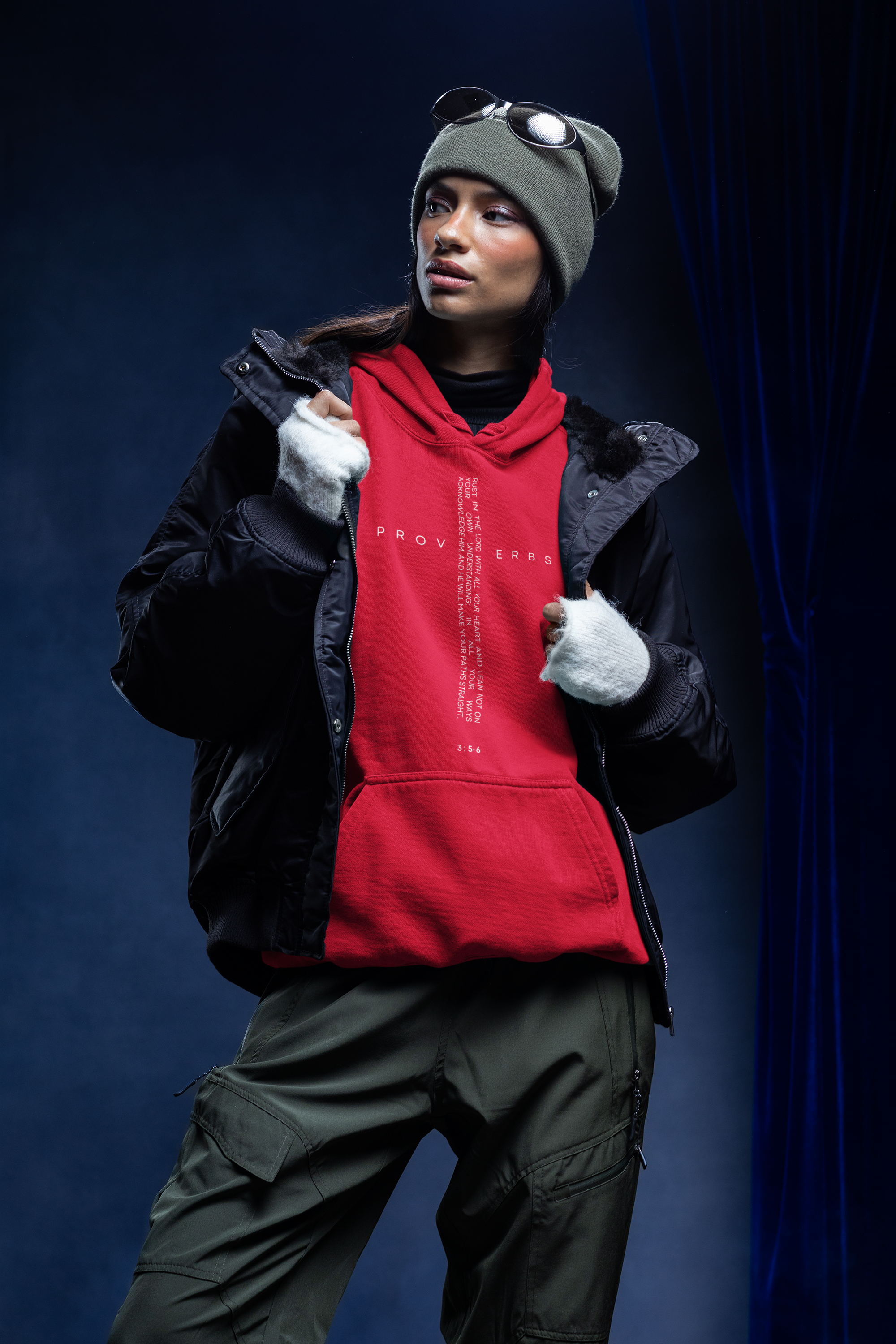 A person dressed in outdoor winter clothing, including a beanie, sunglasses, a black jacket, a red hoodie, and gloves, standing against a dark background.