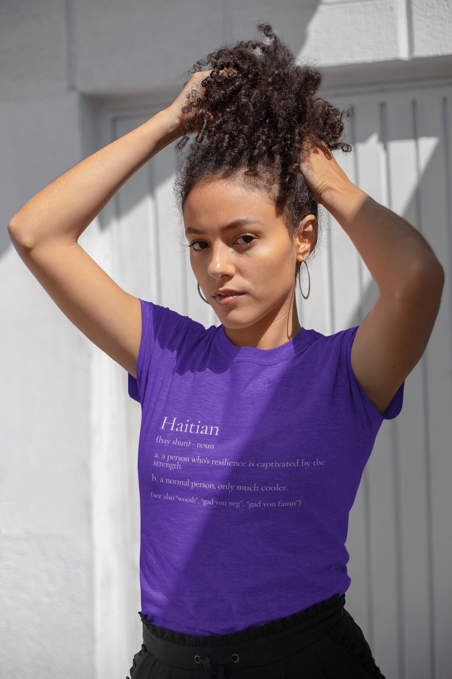A woman with curly hair tied up in a high bun, wearing a purple T-shirt with text about Haitians, standing outdoors in front of a white textured wall.