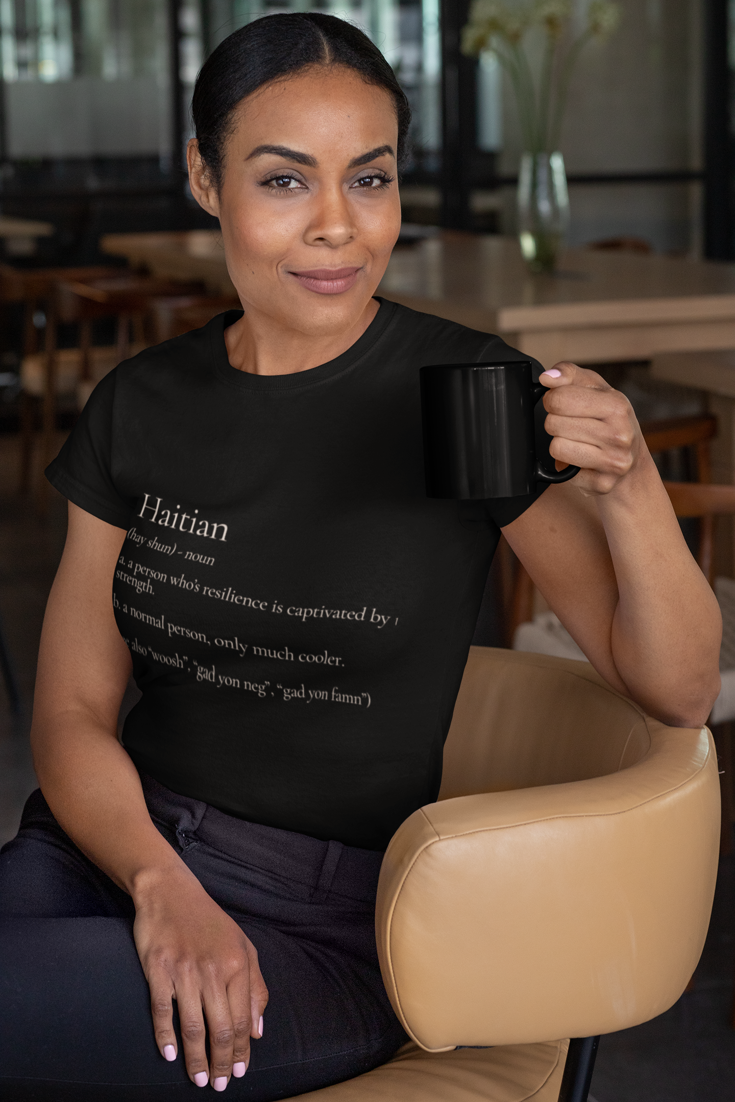 A woman with dark hair and brown skin sitting in a tan chair in a modern room with large windows, holding a black mug, wearing a black T-shirt with text about Haitians.