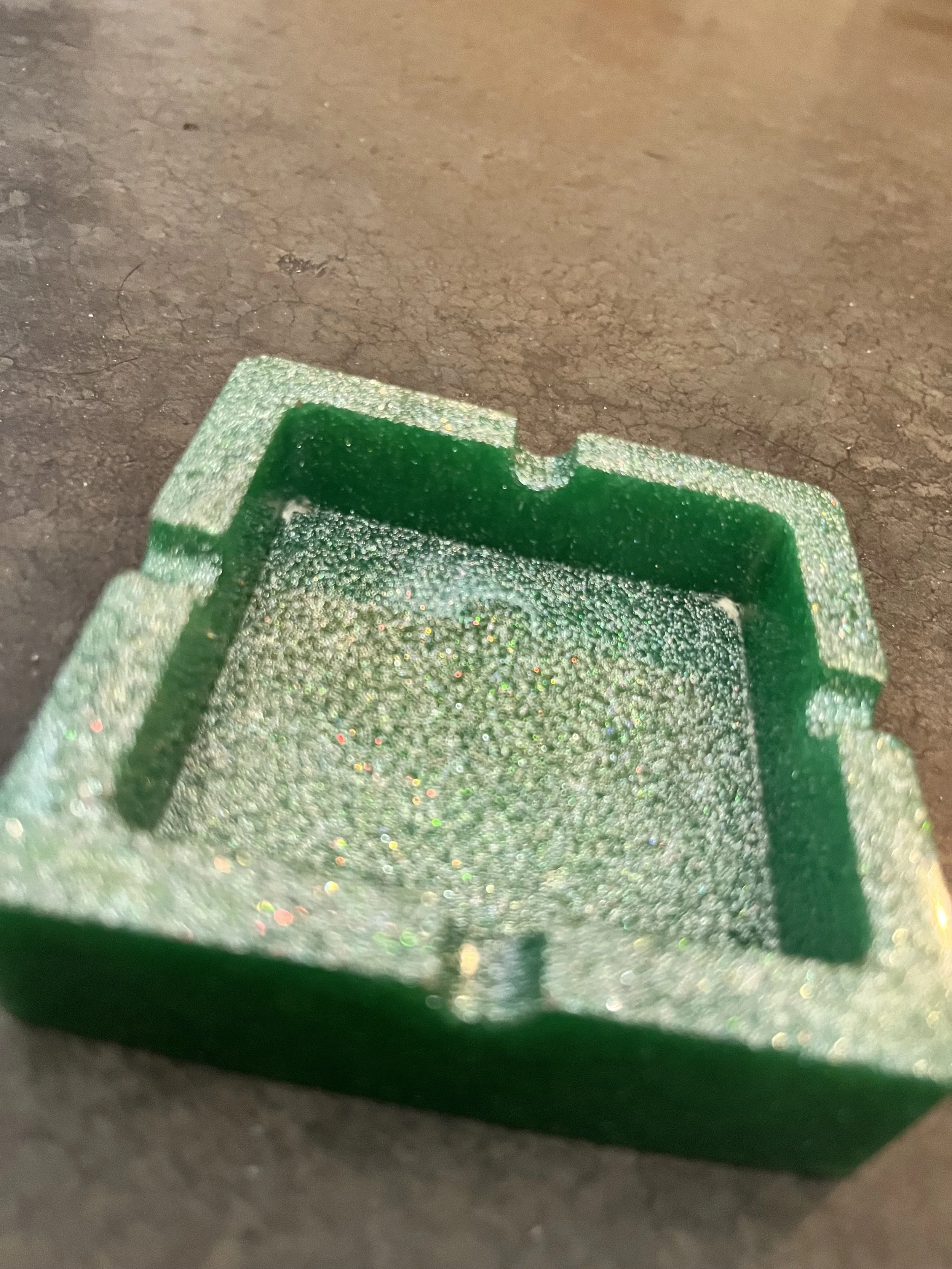 Close-up of a green and silver glittery object with a rectangular slot on a textured brown surface.