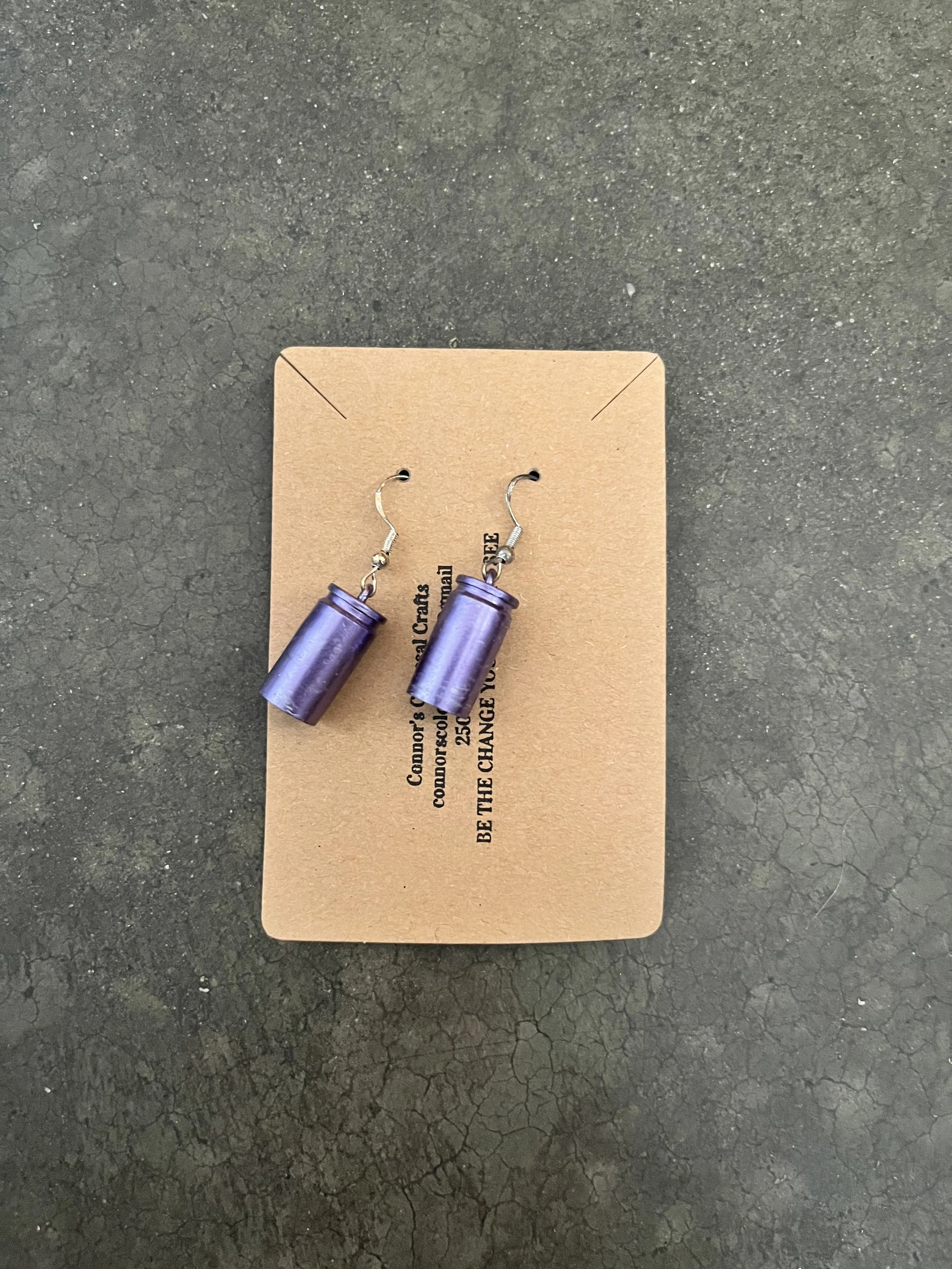 A pair of purple gun casing earrings on a brown card placed on a cracked black surface.