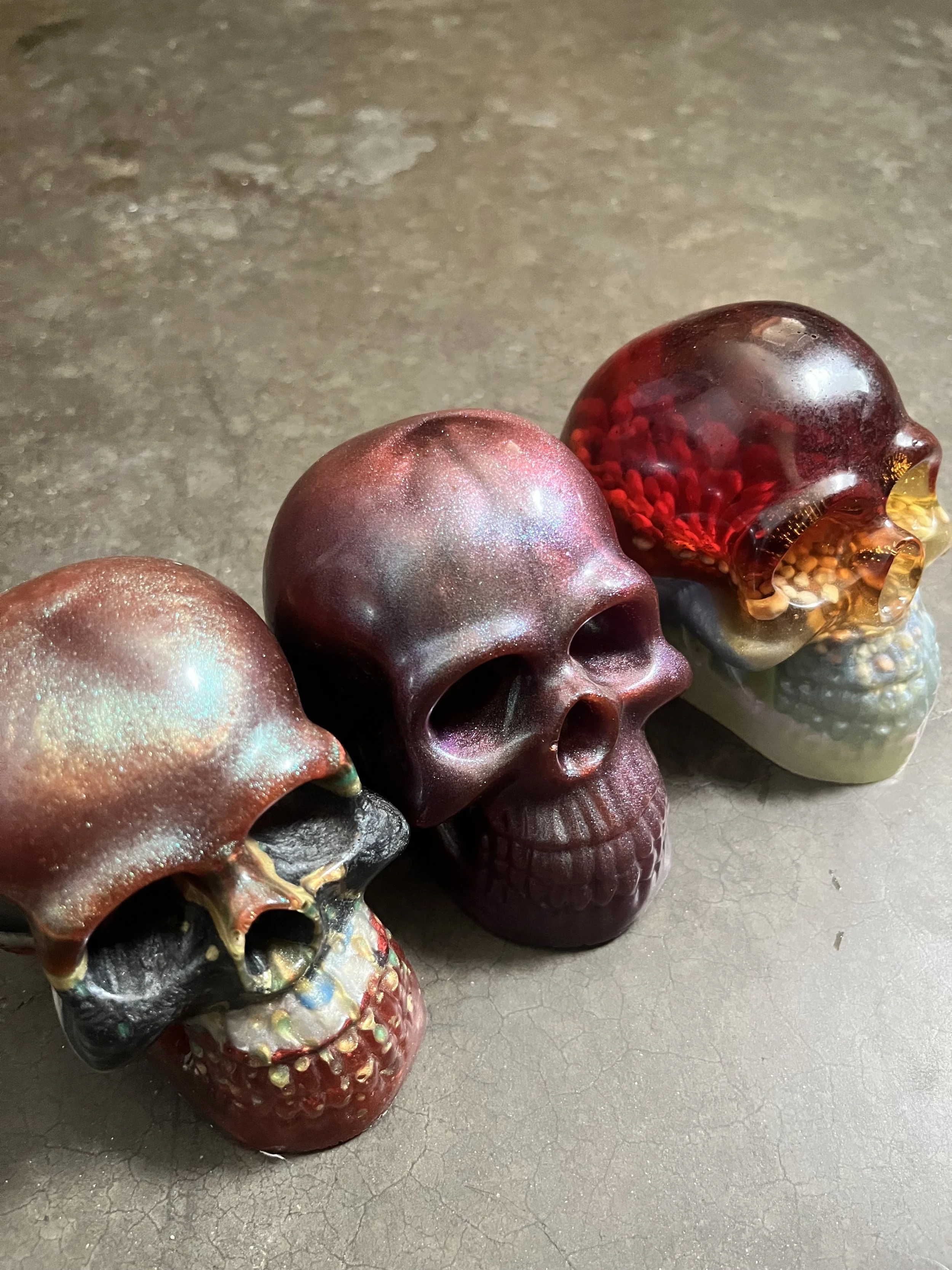 A row of handmade decorative skulls with a shiny, metallic finish, featuring various colors such as red, gold, and silver, placed on a gray, textured surface.