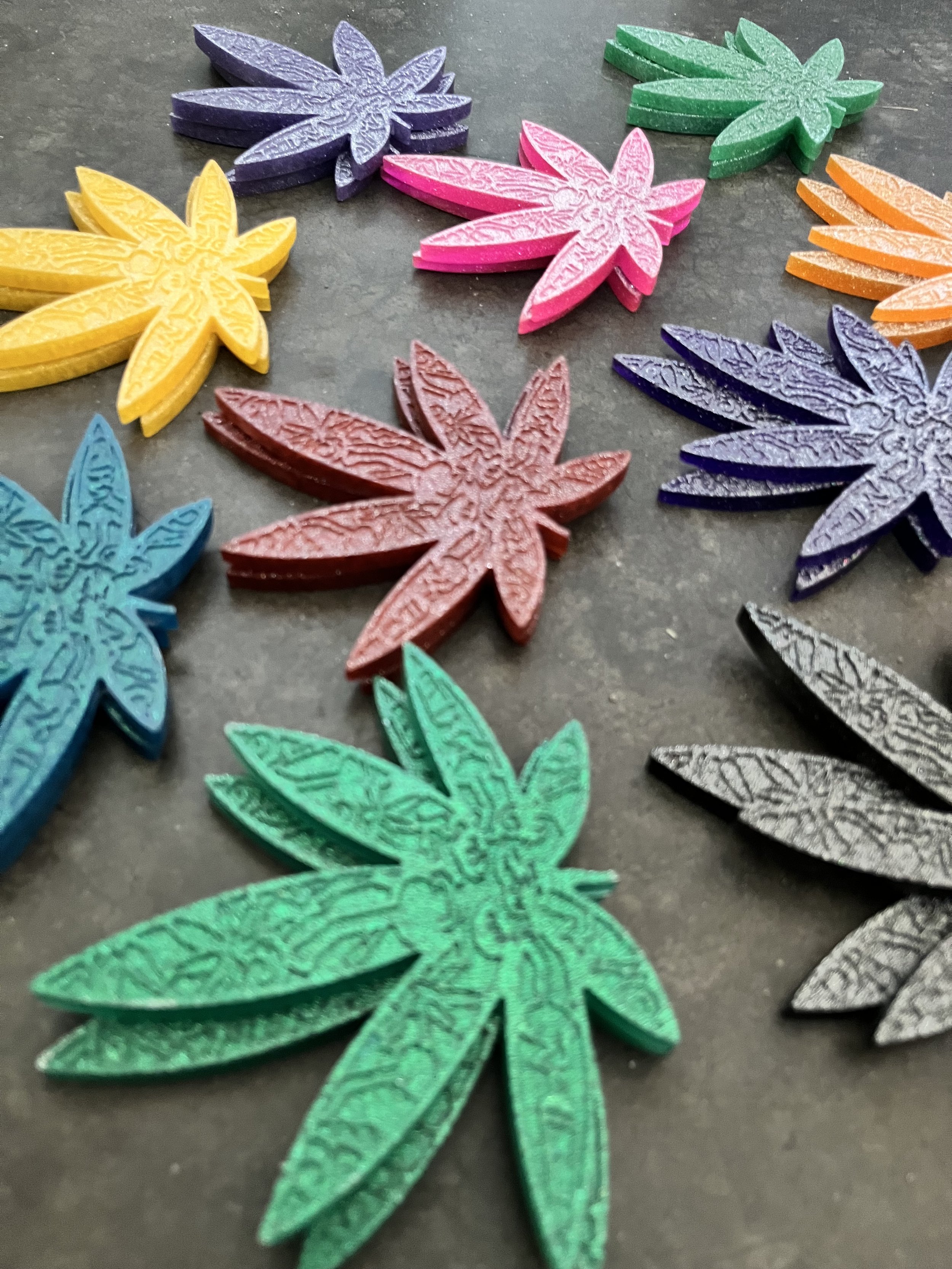 Colorful handmade marijuana leaf coasters in yellow, purple, pink, teal, orange, and black laid out on a dark surface.
