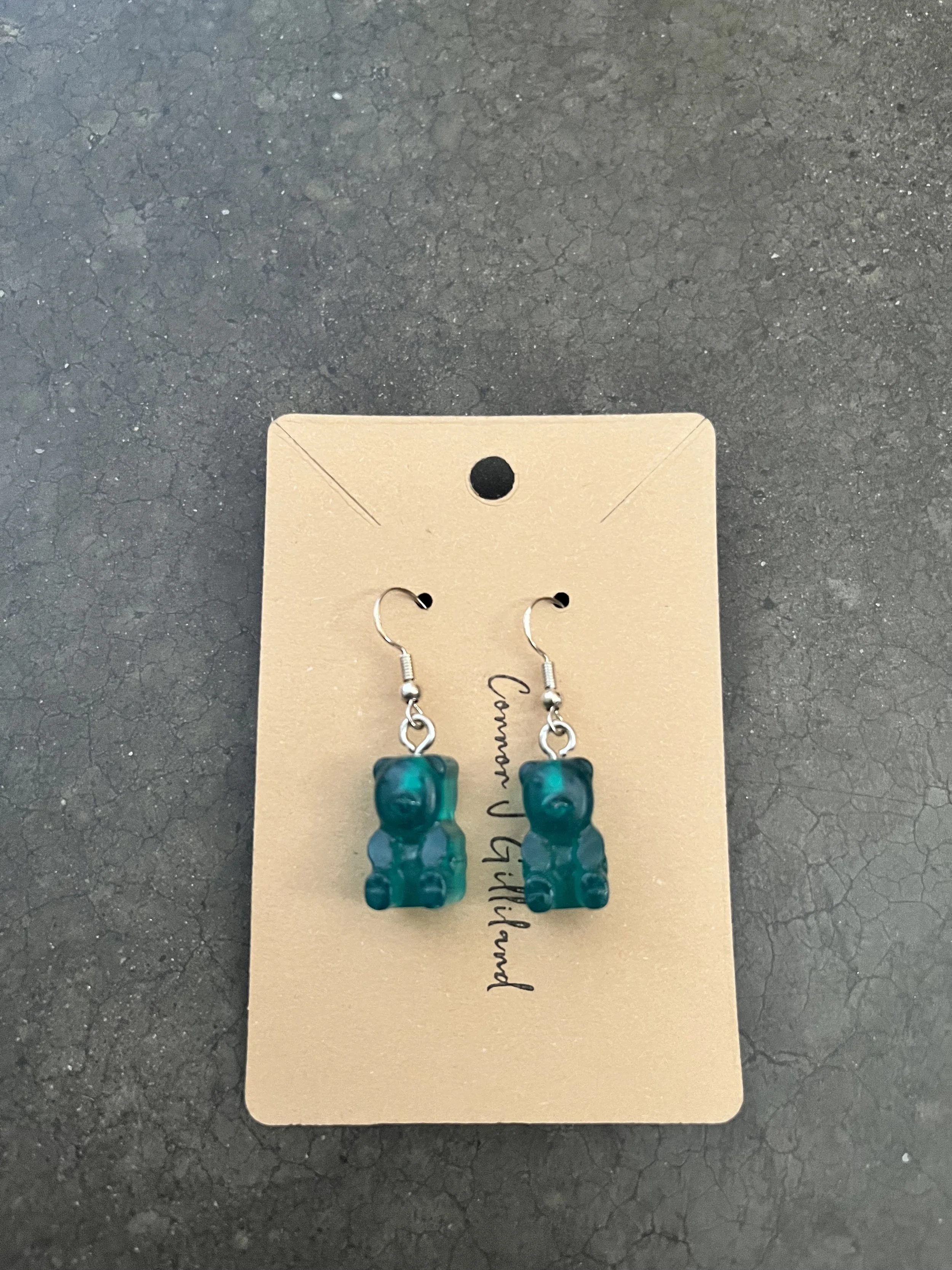 Pair of turquoise bear-shaped earrings hanging from a beige card with handwritten-style text, on a dark textured surface.