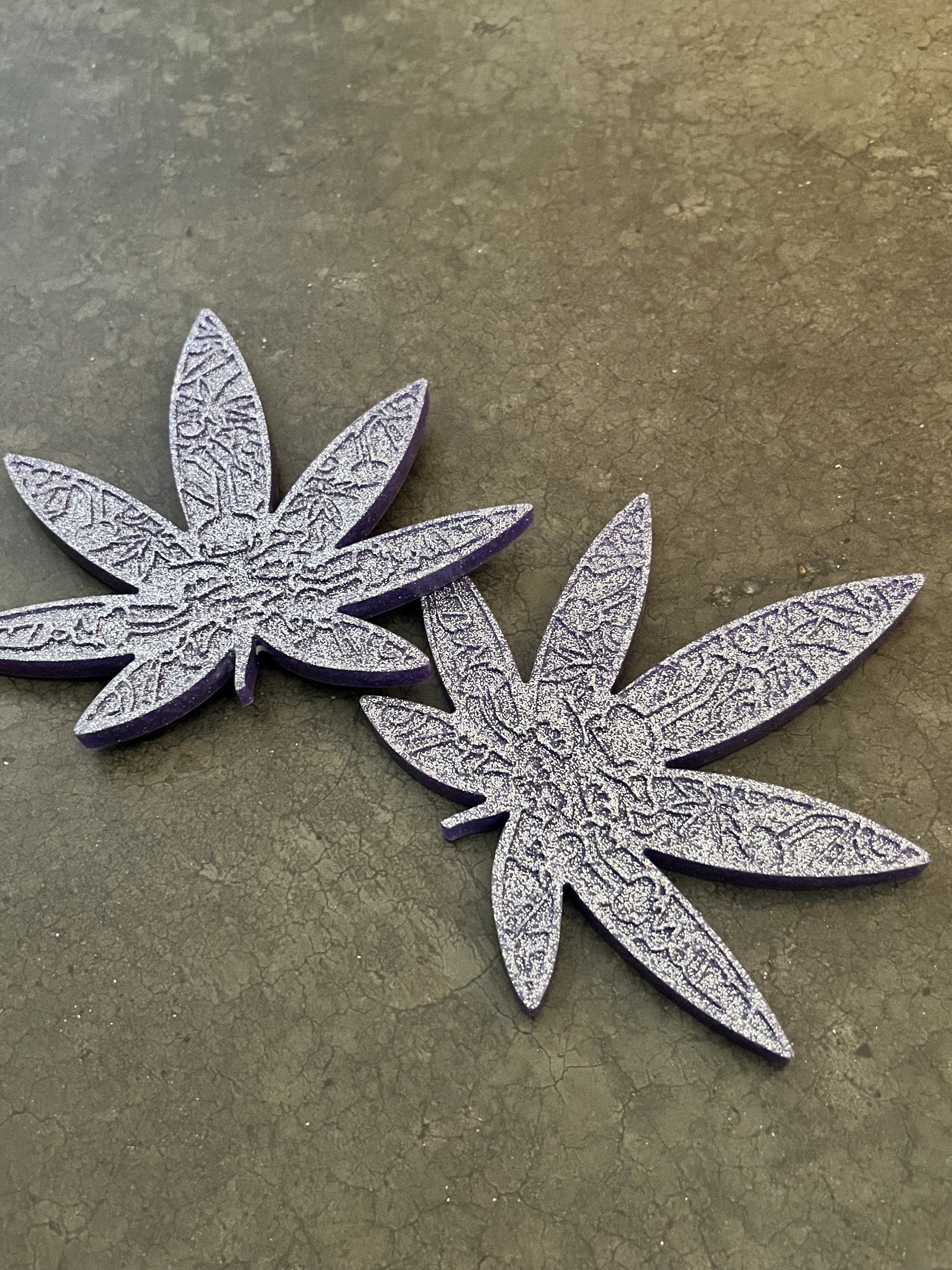 Decorative glittery silver and purple cannabis leaf-shaped coasters on a cracked dark gray surface.
