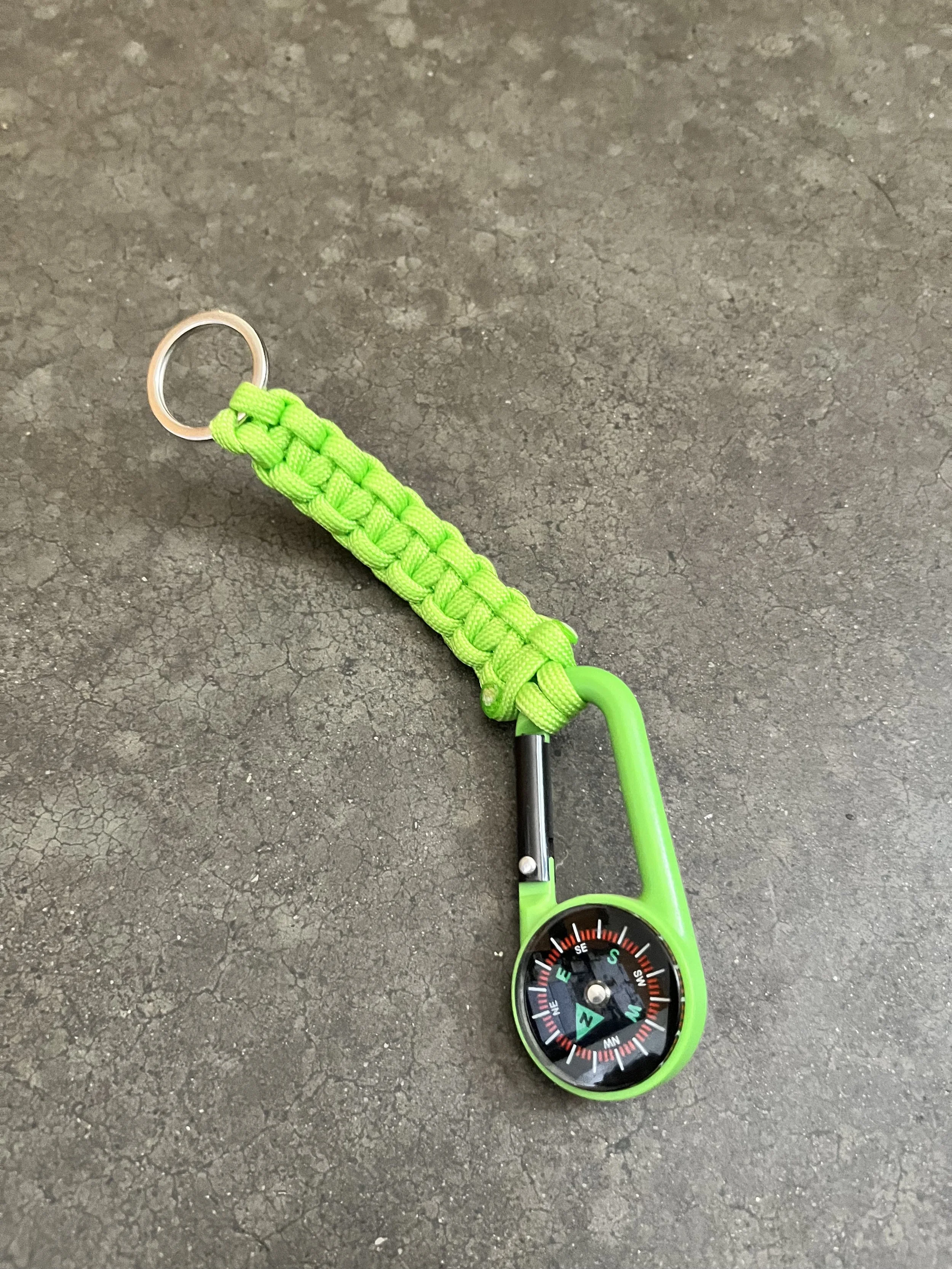 A green compass keychain with a braided paracord strap, shown on a rough textured gray surface.