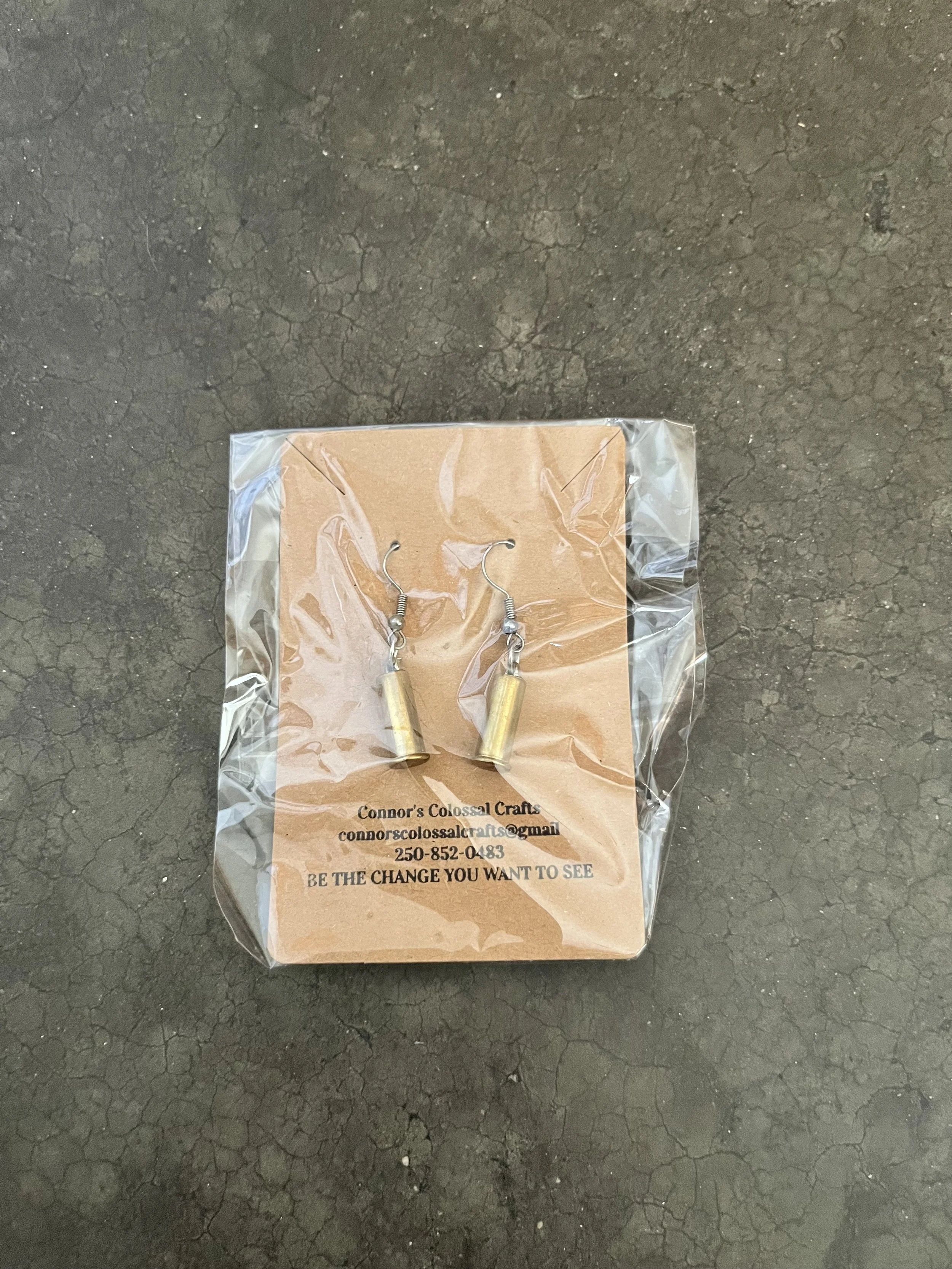 A pair of gun casing earrings in a plastic bag attached to a cardboard backing with contact information and a message, on a dark, cracked surface.