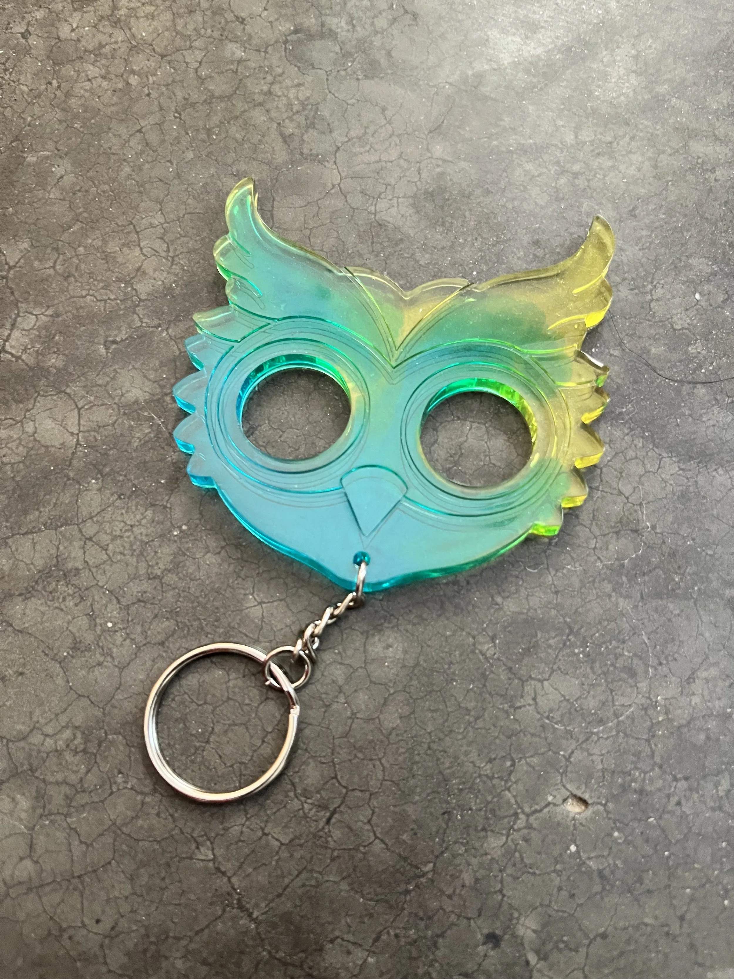A rainbow-colored, wing-shaped epoxy resin keychain with two eye holes, attached to a metal keyring, lying on a cracked black surface.