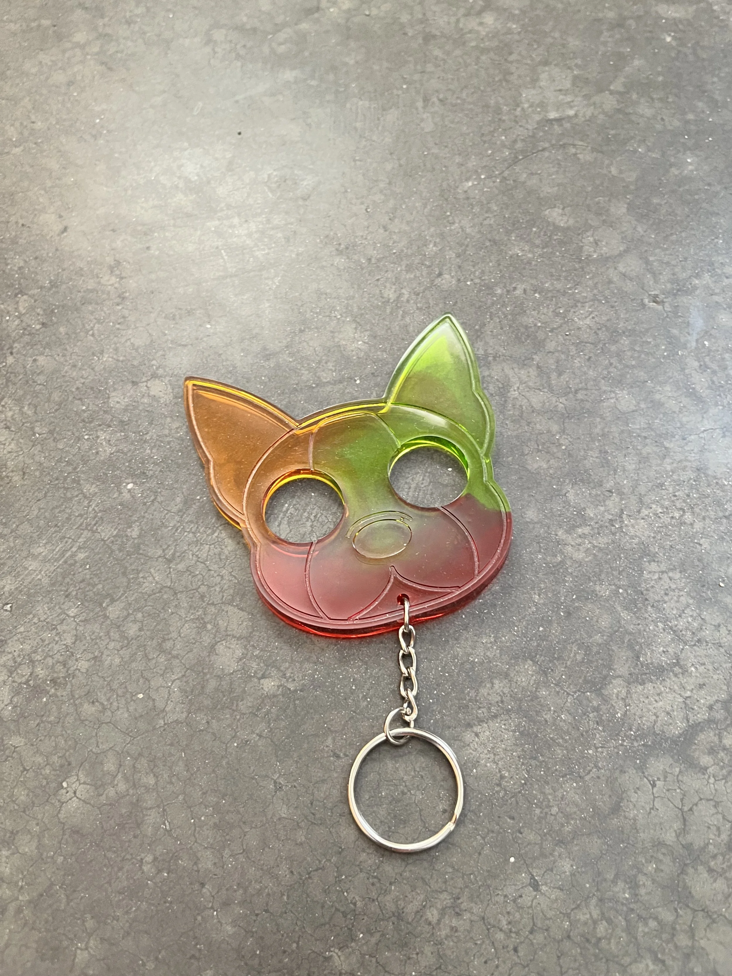 A colorful cat-shaped keychain made of translucent plastic, with a gradient of red, orange, yellow, green, and red, attached to a metal keyring and chain, lying on a black textured surface.