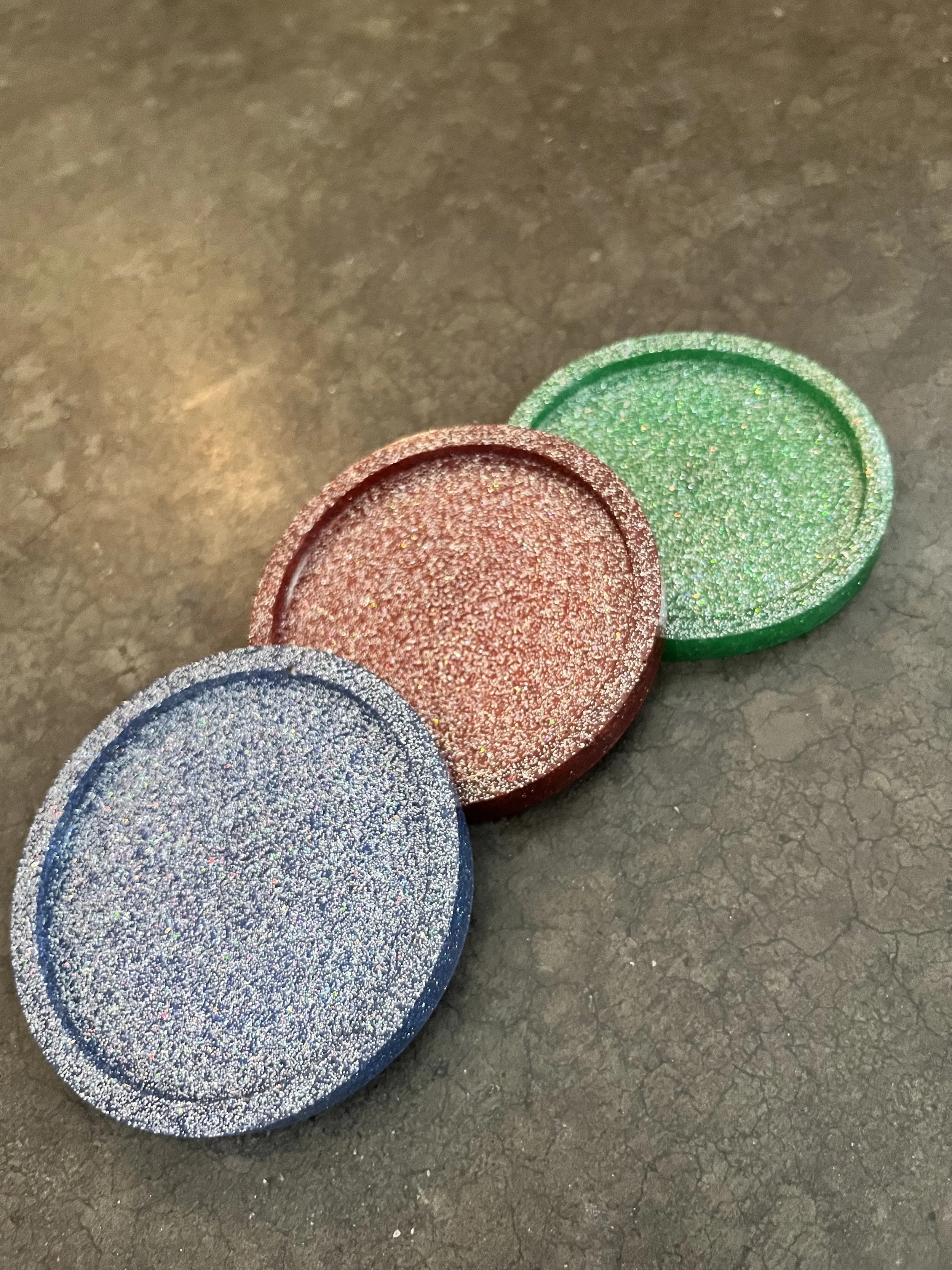 Three round makeup containers with glittery lids in blue, red, and green, placed on a dark textured surface.