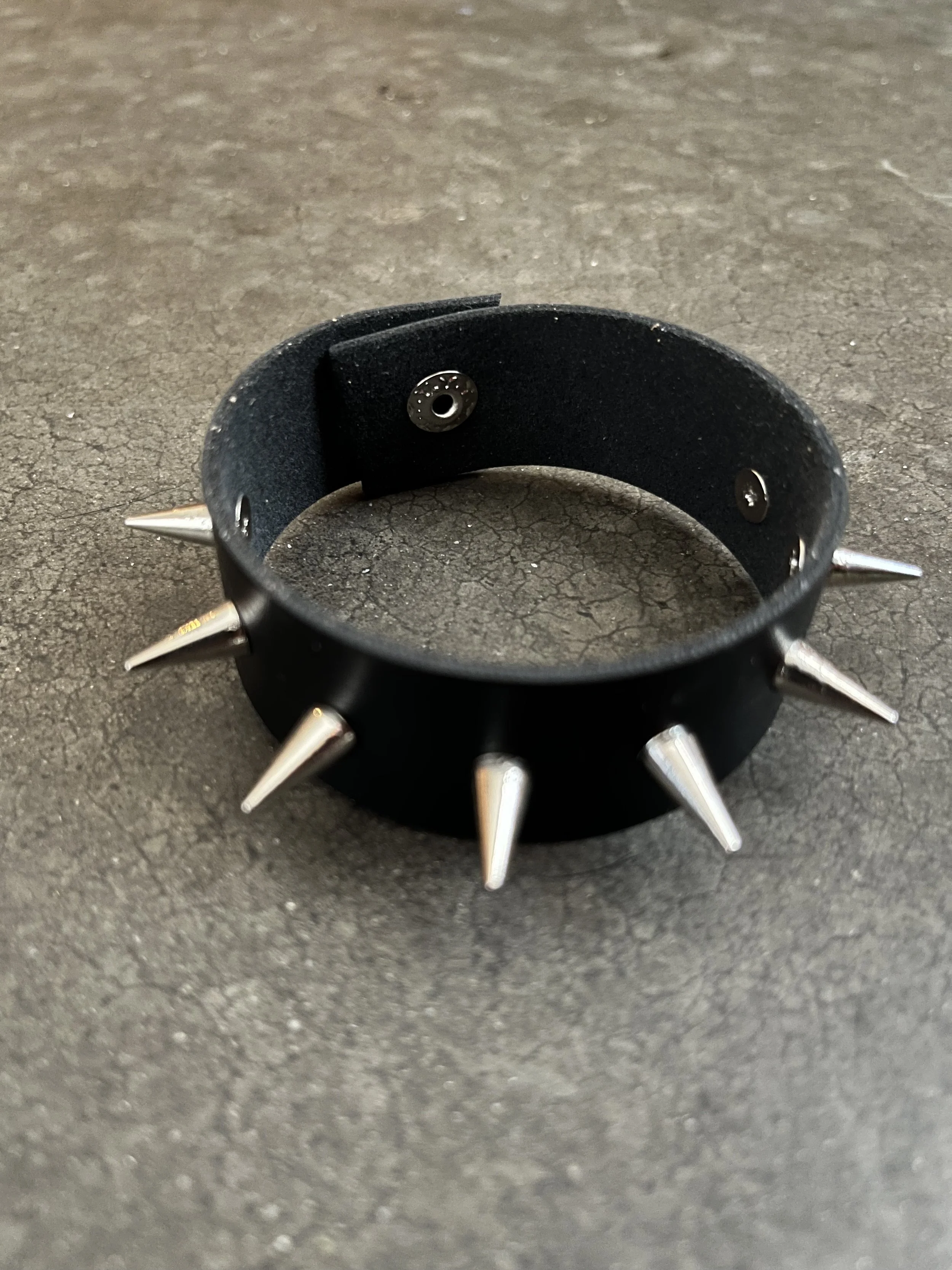 A black leather wristband with metal spikes and snaps, placed on a rough black surface.