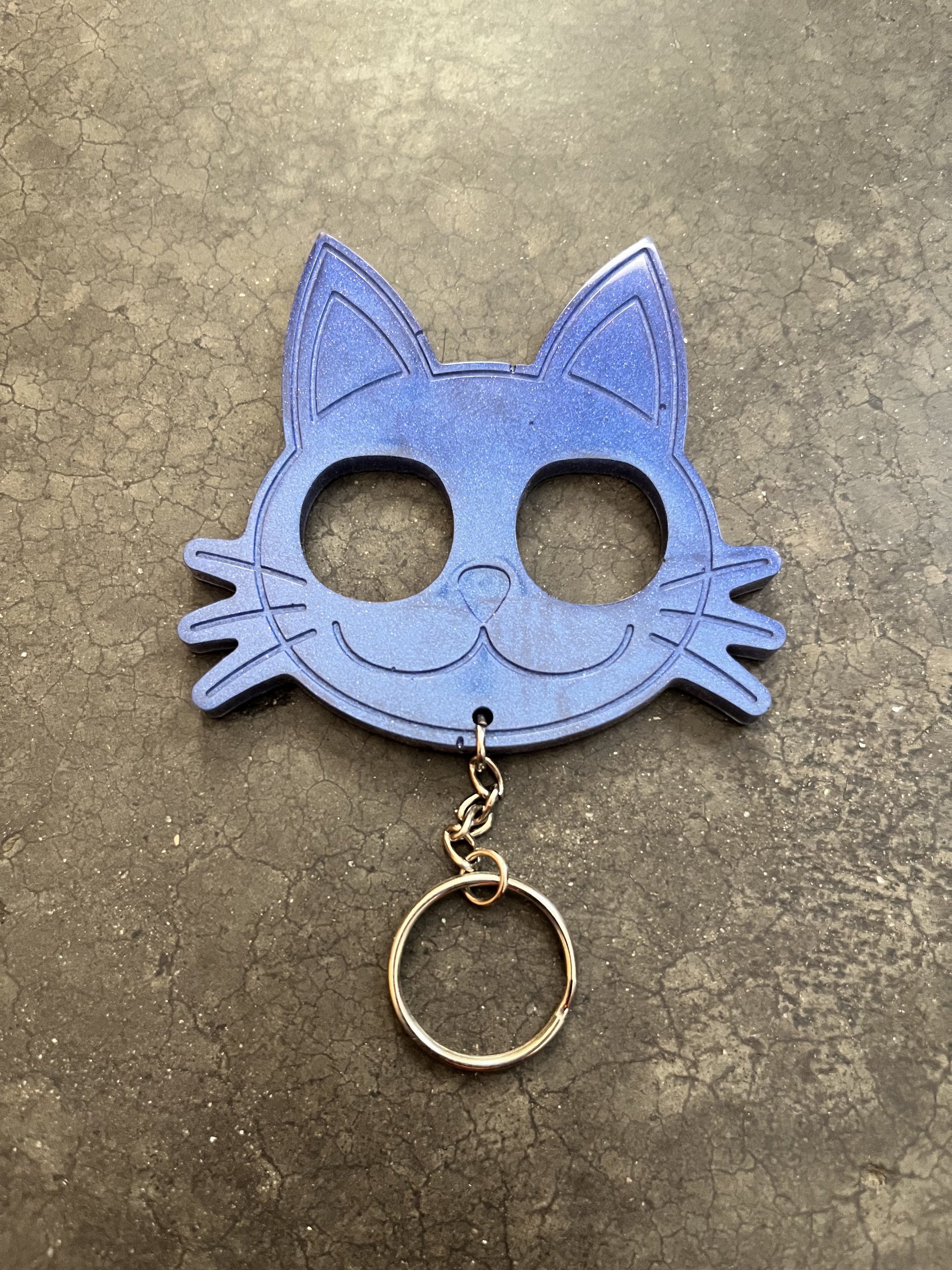 A keychain shaped like a blue cat's face with cutout eyes, nose, and whiskers, attached to a silver ring, on a textured dark surface.