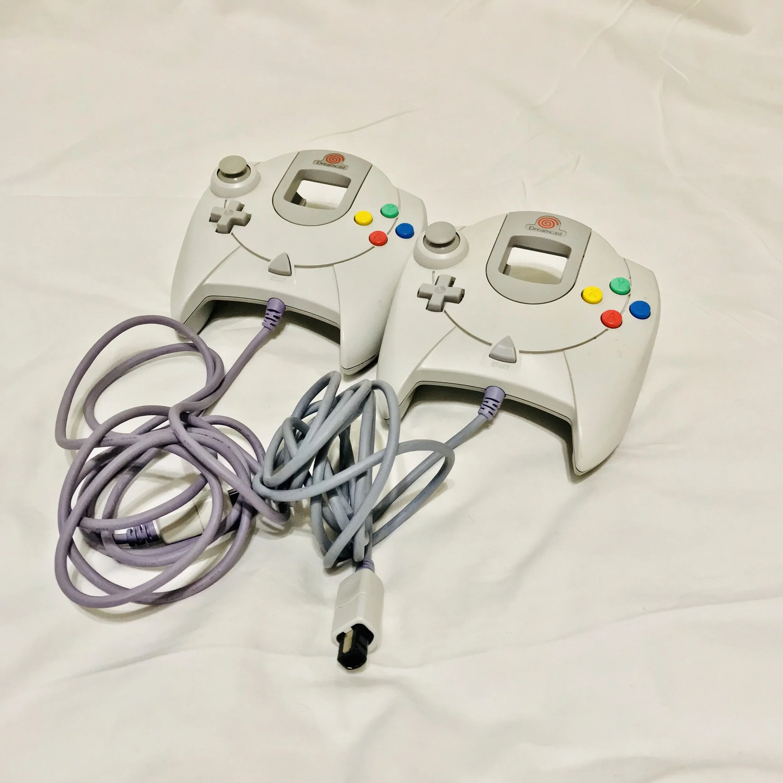 Two white Sega Dreamcast controllers connected with cords on a white surface.