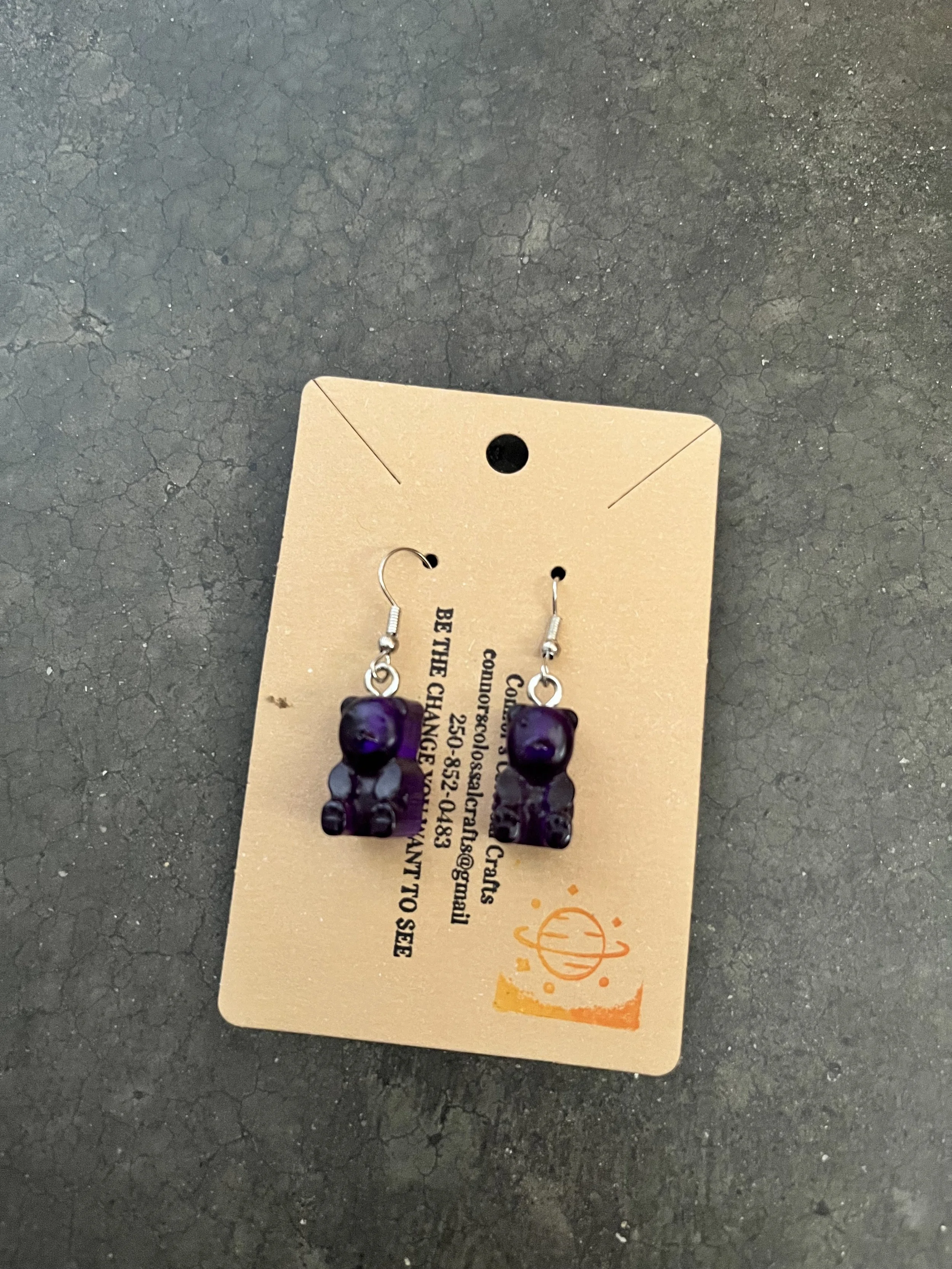 Pair of gummy bear earrings on a brown paper card, on a dark textured surface.