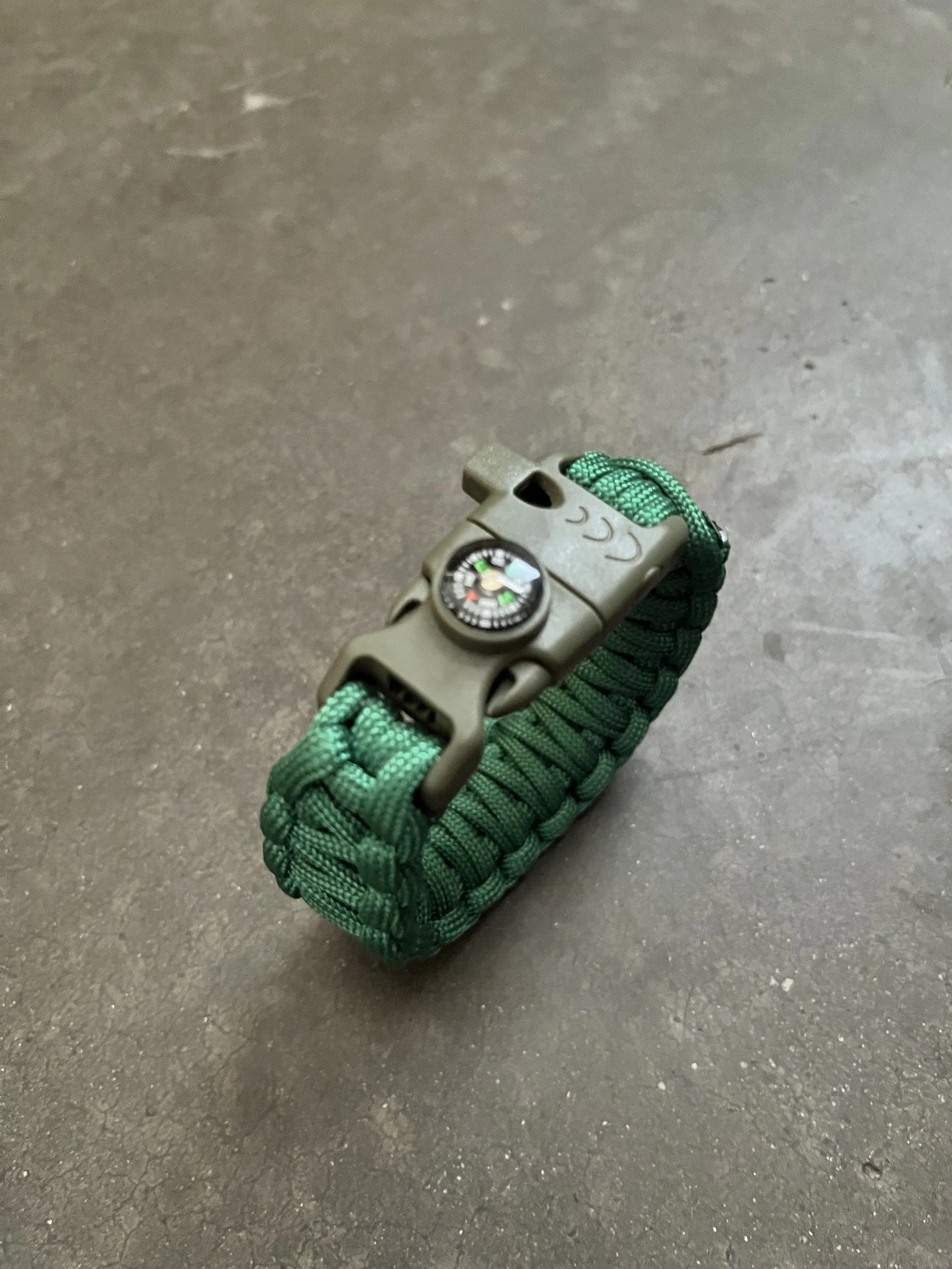 A green paracord bracelet with a compass attached, worn on a person's wrist.