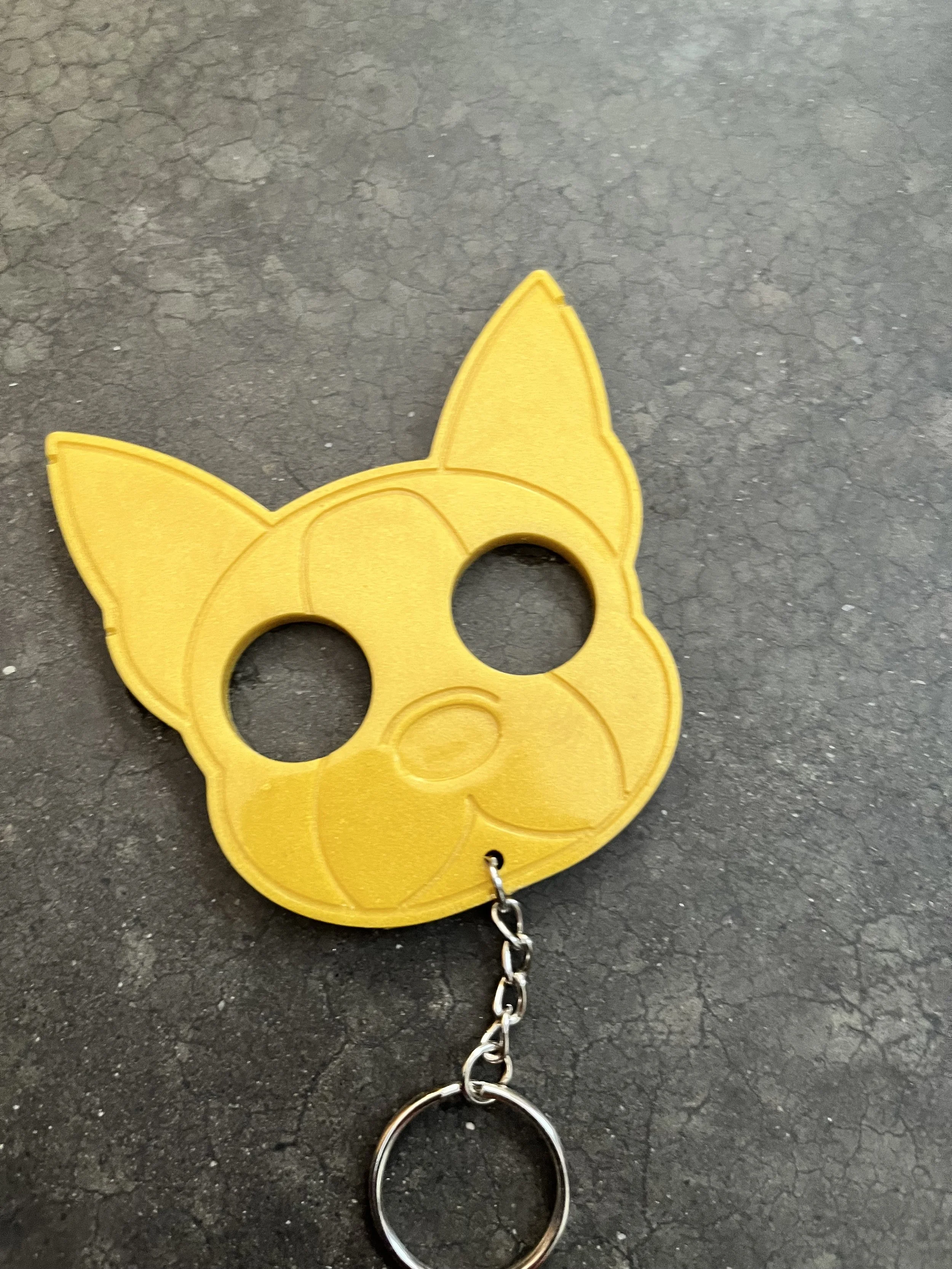Yellow keychain in the shape of a cat's head with pointed ears and two circular holes, attached to a metal chain, lying on a dark, cracked surface.