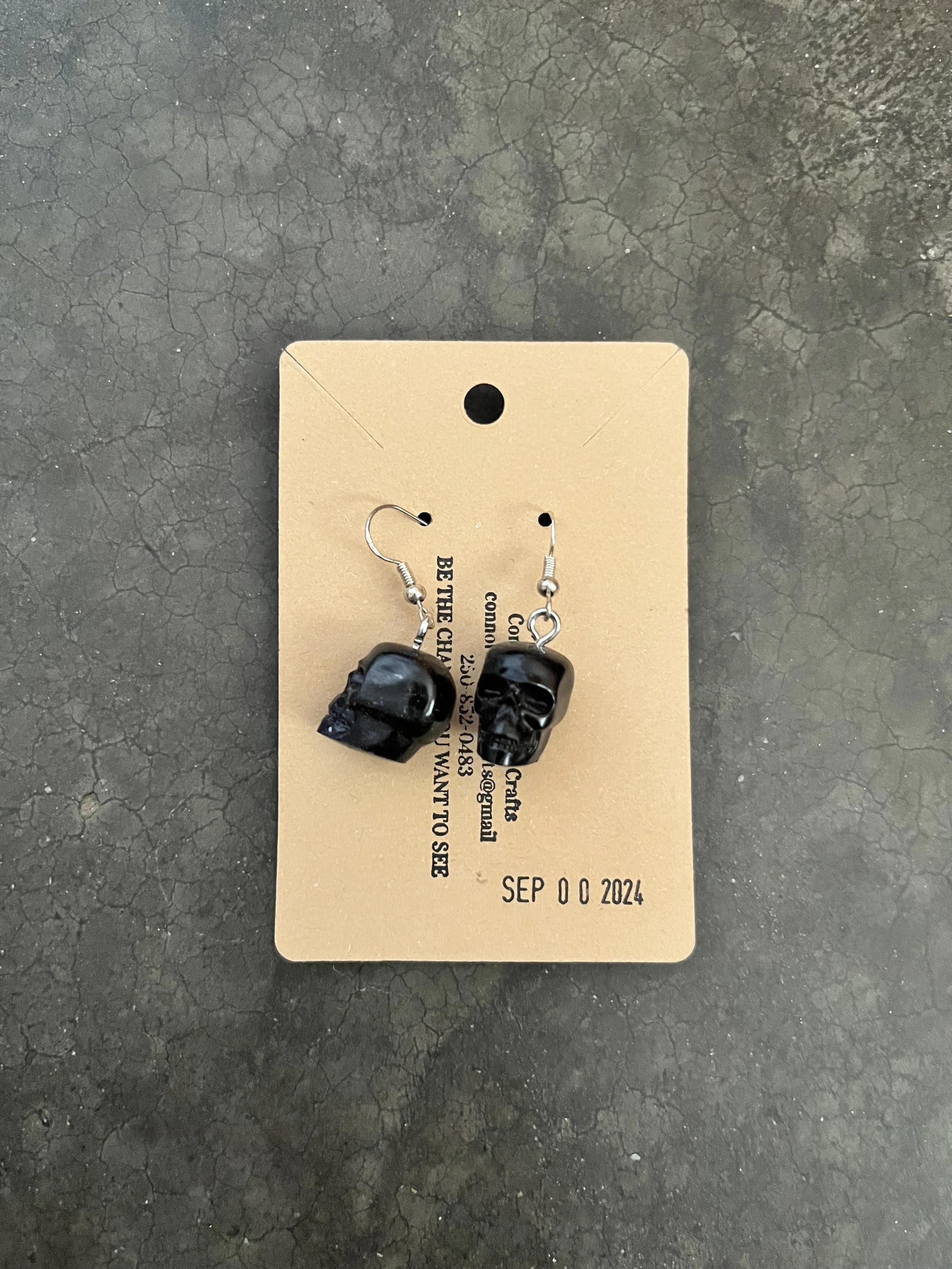 Black epoxy resin earrings,attached to a brown rectangular paper tag with text, on a black cracked surface.