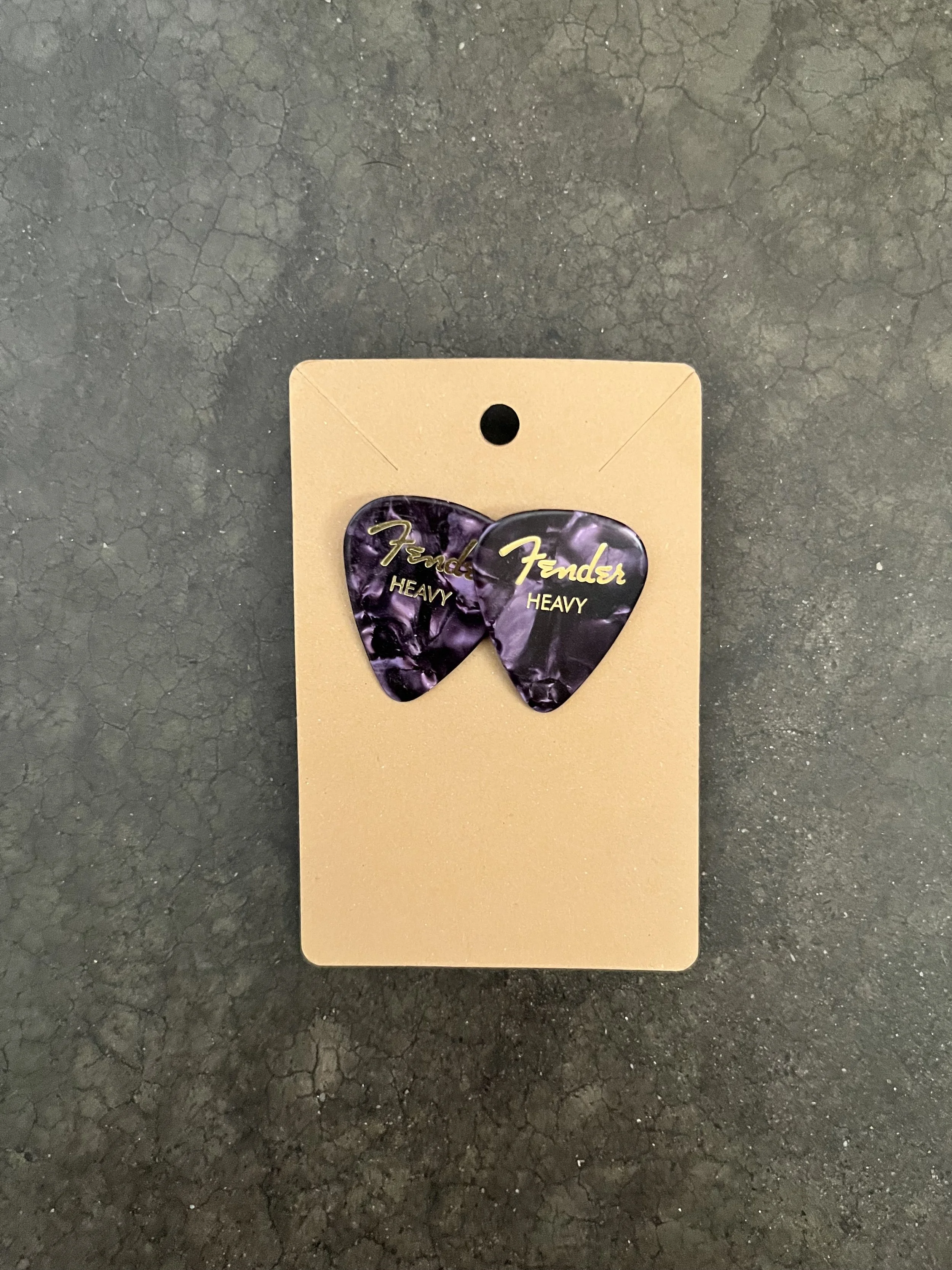 A beige card on a black textured surface with two purple guitar picks attached, each labeled 'Fender HEAVY' in gold text.Earrings that are handmade.