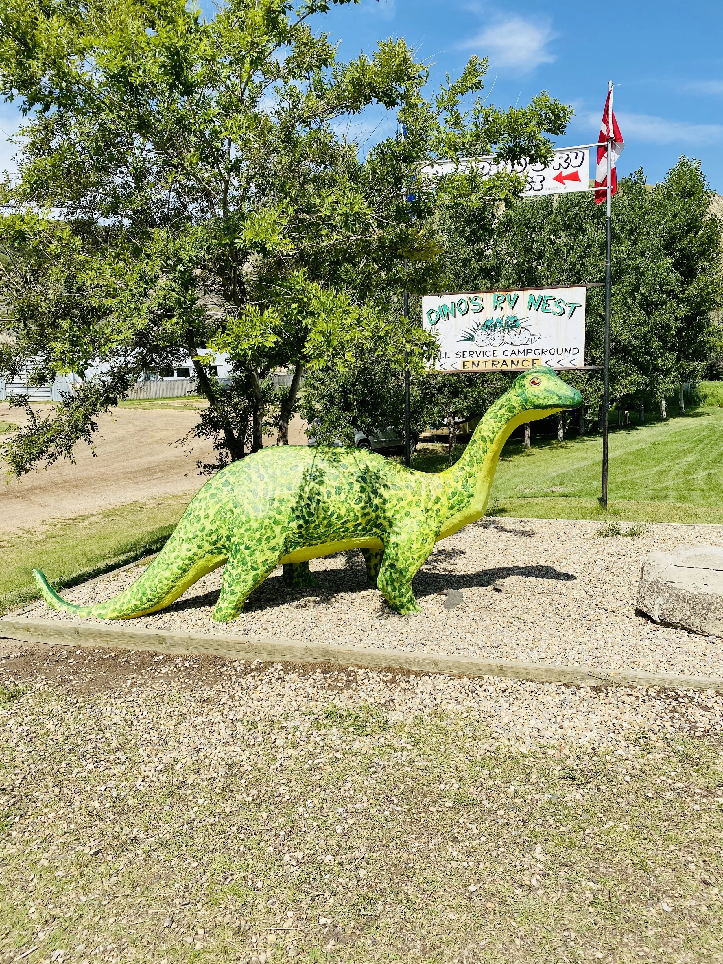 A large, green, dinosaur-shaped sculpture in a park with trees and a dirt path. Signage in the background reads "Dino's RV Nest" and indicates an entrance, with flags and a blue sky overhead.