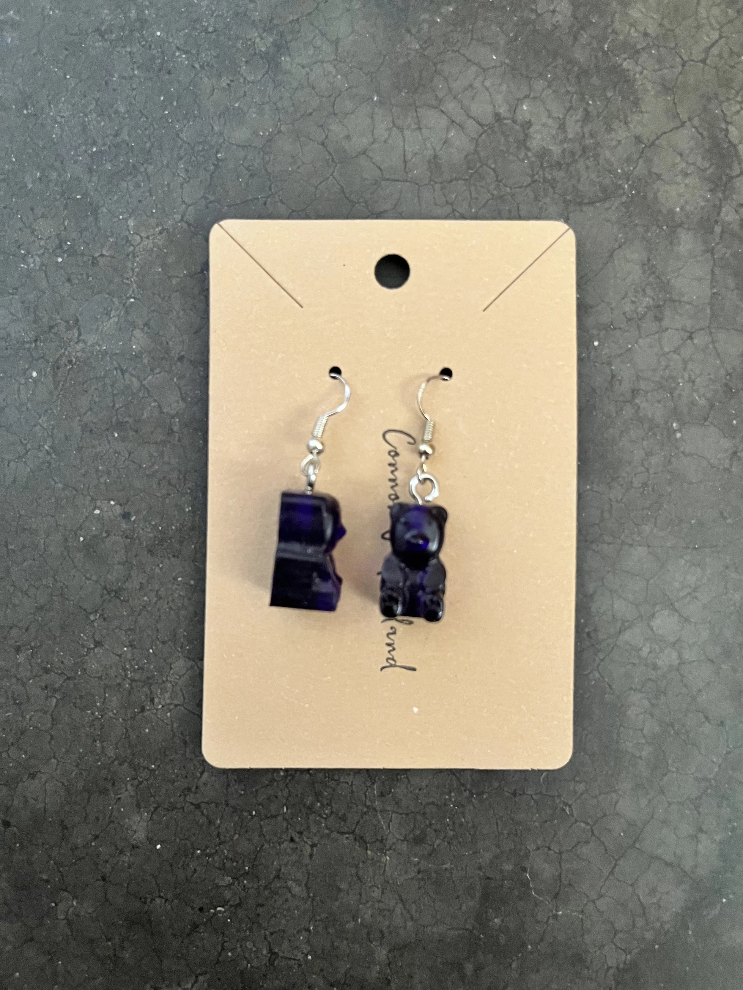 A handmade pair of black gummy bear earrings displayed on a beige card.