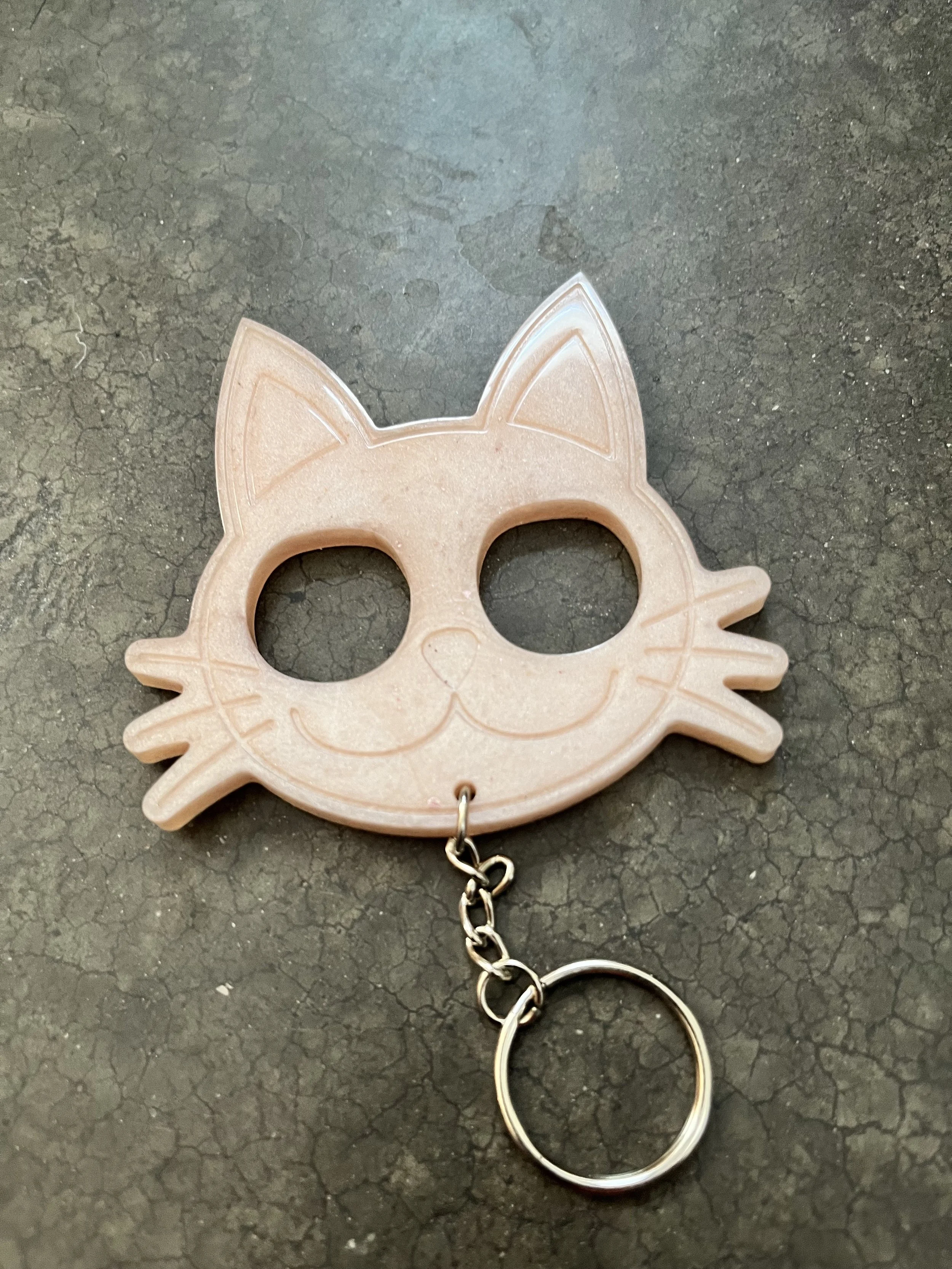 A pink cat-shaped keychain with large round eyes and a small tail, attached to a silver chain and keyring, on a dark, textured surface.