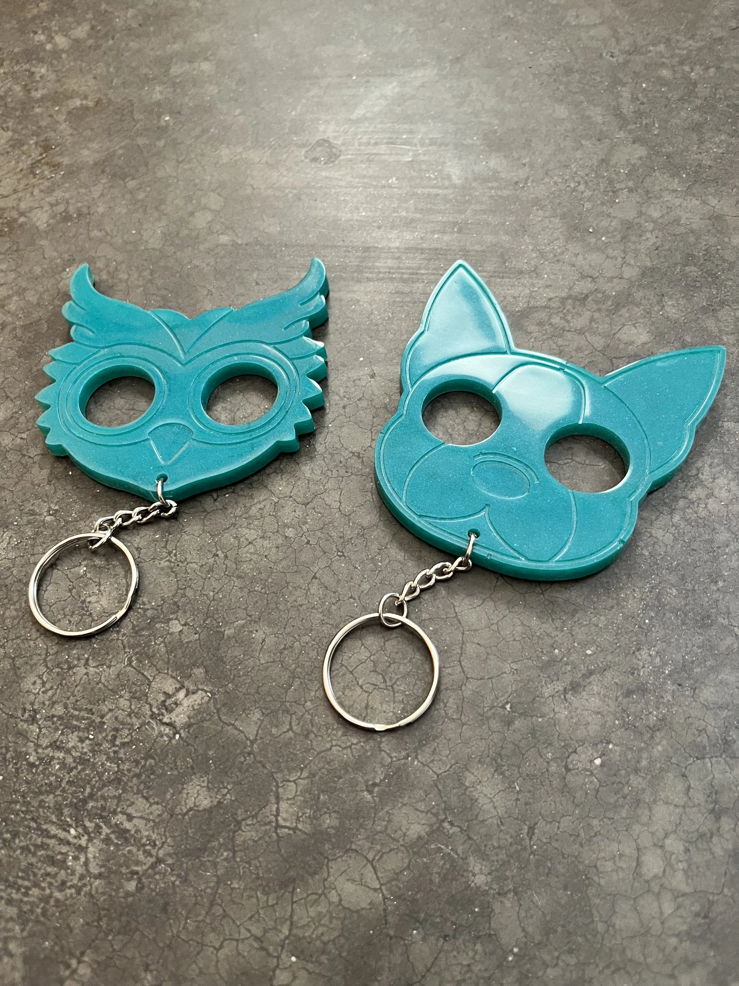 Two teal-colored handmade epoxy resin emergency keychains, on a dark cracked surface.