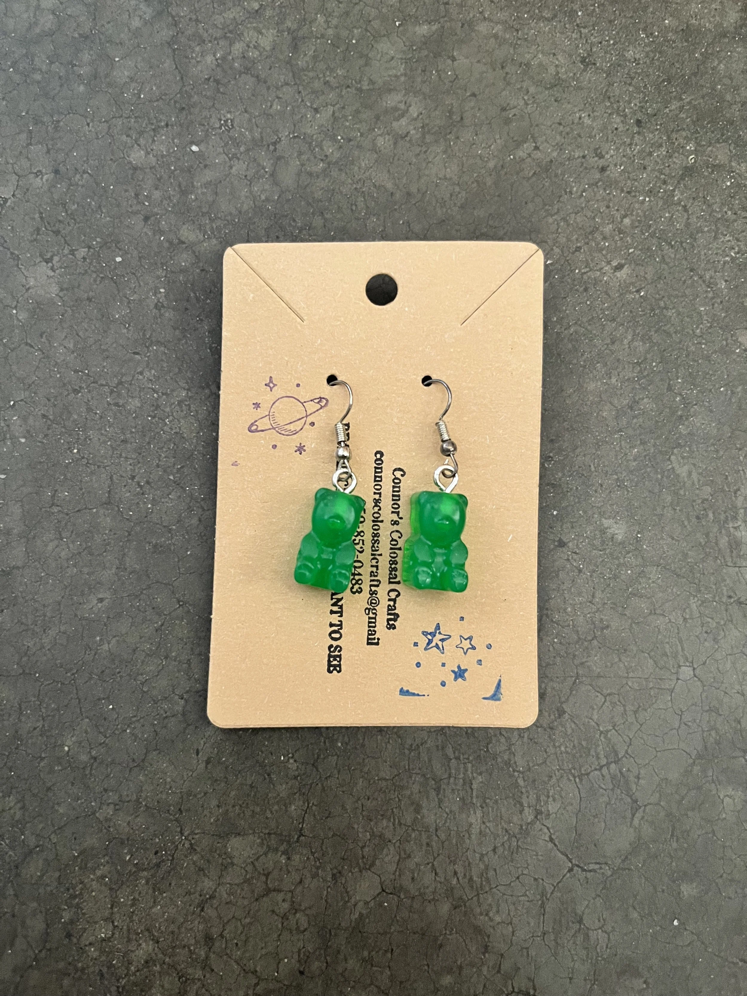 Green gummy bear earrings on a brown card, placed on a black textured surface.