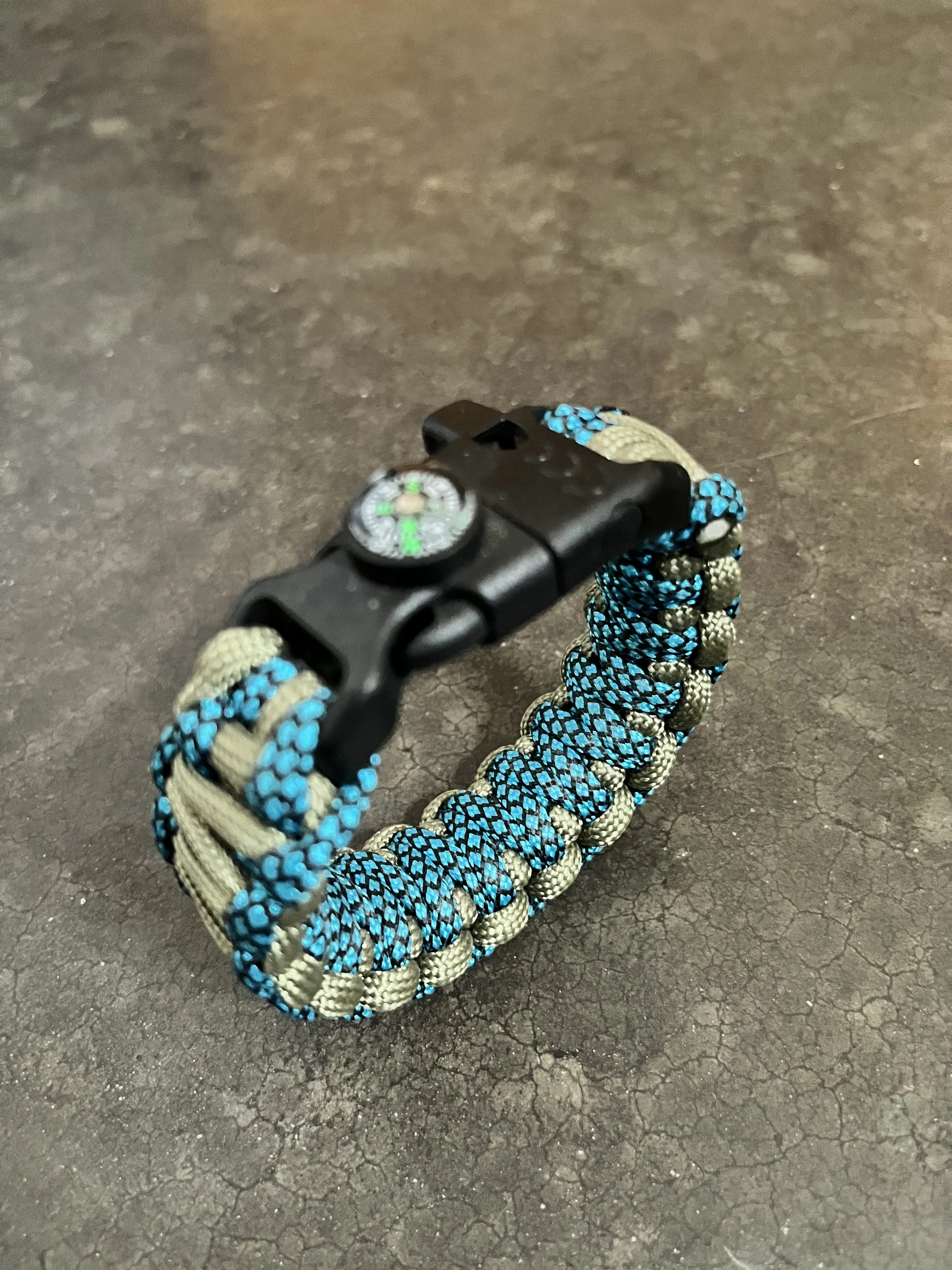 Close-up of a paracord bracelet with a black metal clasp and a compass attached, resting on a cracked asphalt surface.