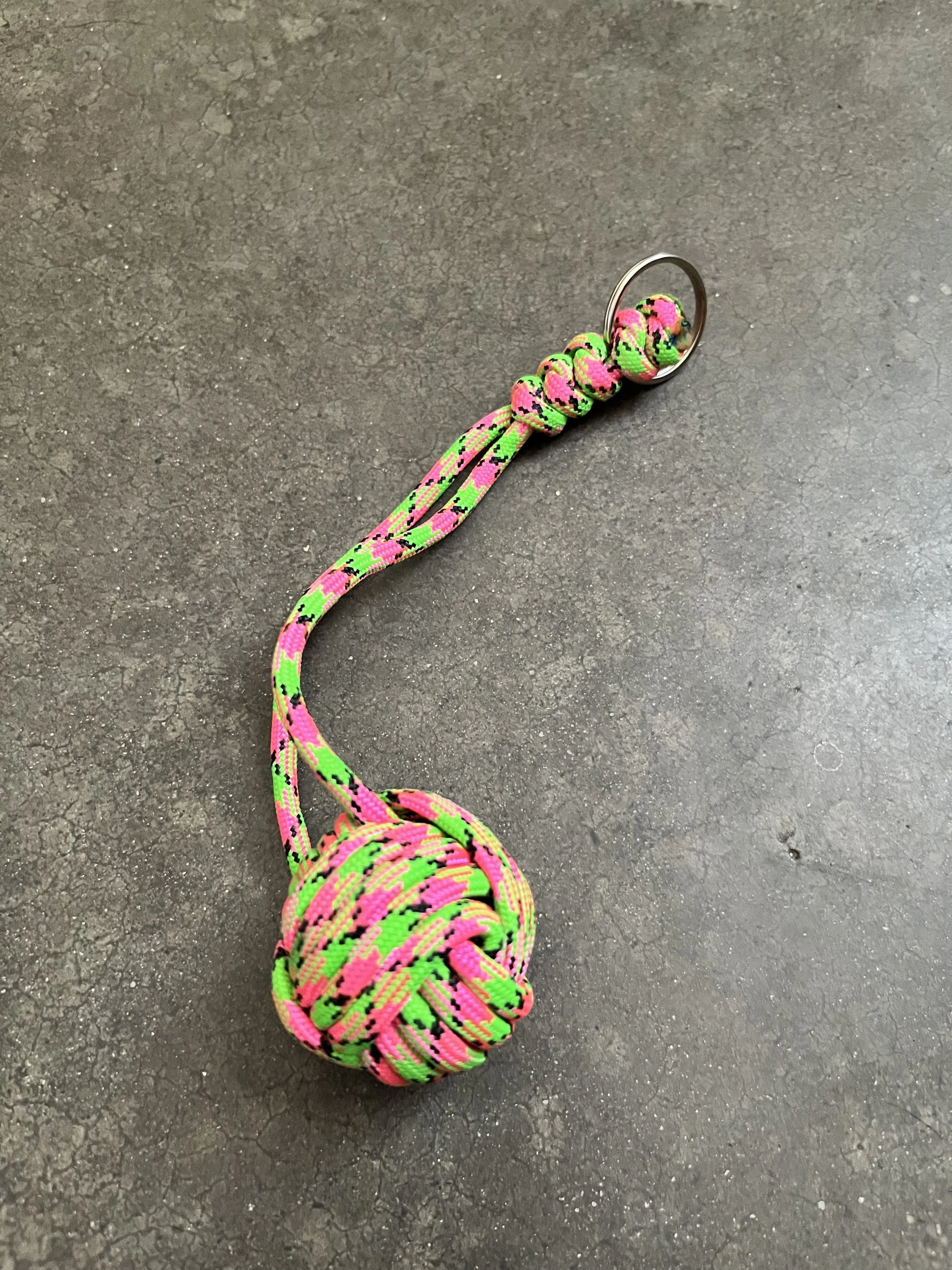Colorful neon pink and green paracord emergency ball keychain, on a dark textured surface.
