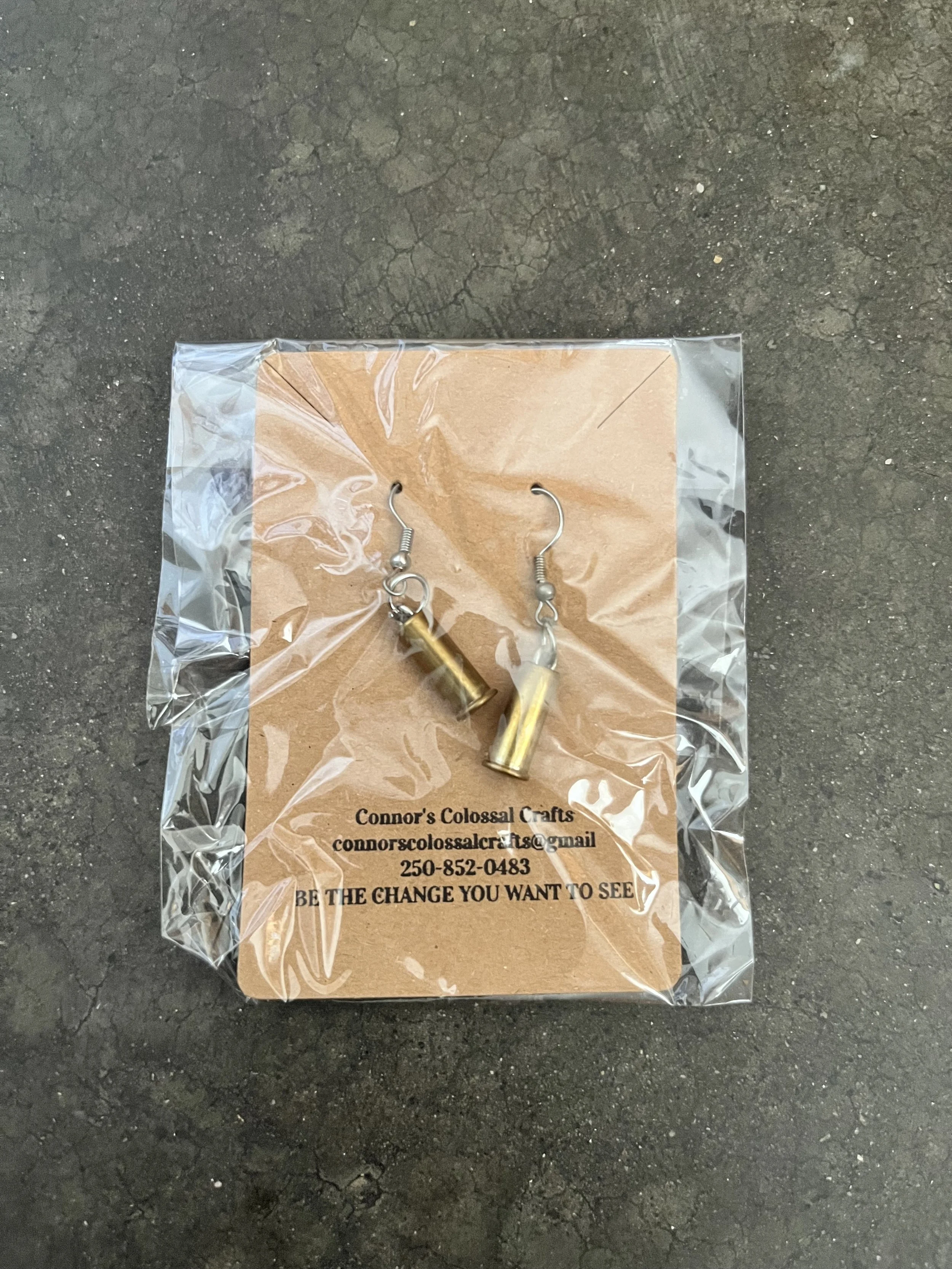 A pair of gun casing earrings displayed on a small piece of cardboard, wrapped in clear plastic, with contact information and a motivational quote printed on the cardboard, placed on a cracked asphalt surface.
